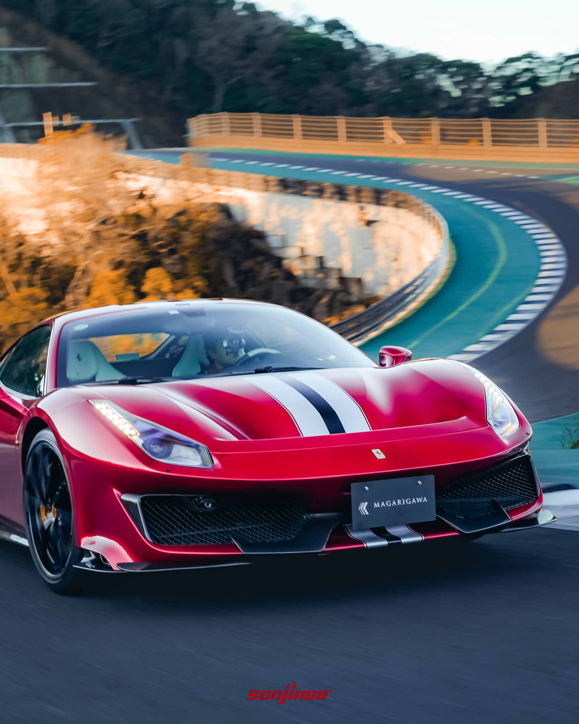 紅色 Ferrari 488 Pista 正駛過帶有紅綠白相間路緣石的彎道，車頭迎向陽光，背景為彎曲的賽道與山壁。