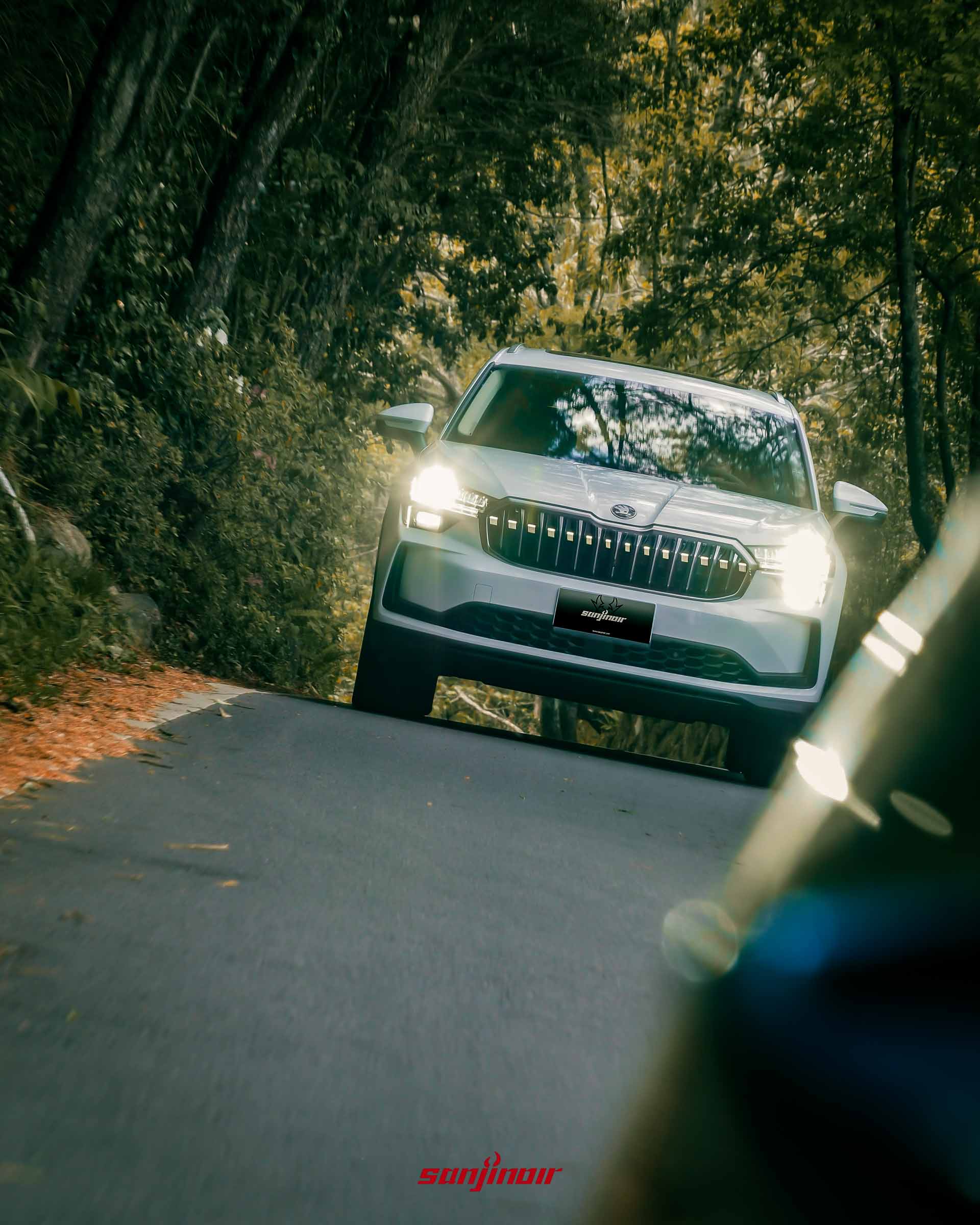 白色 Škoda Kodiaq 2.0 TSI 4×4 行駛在兩側長滿樹木的山區道路上，右下角有模糊的前景物體。