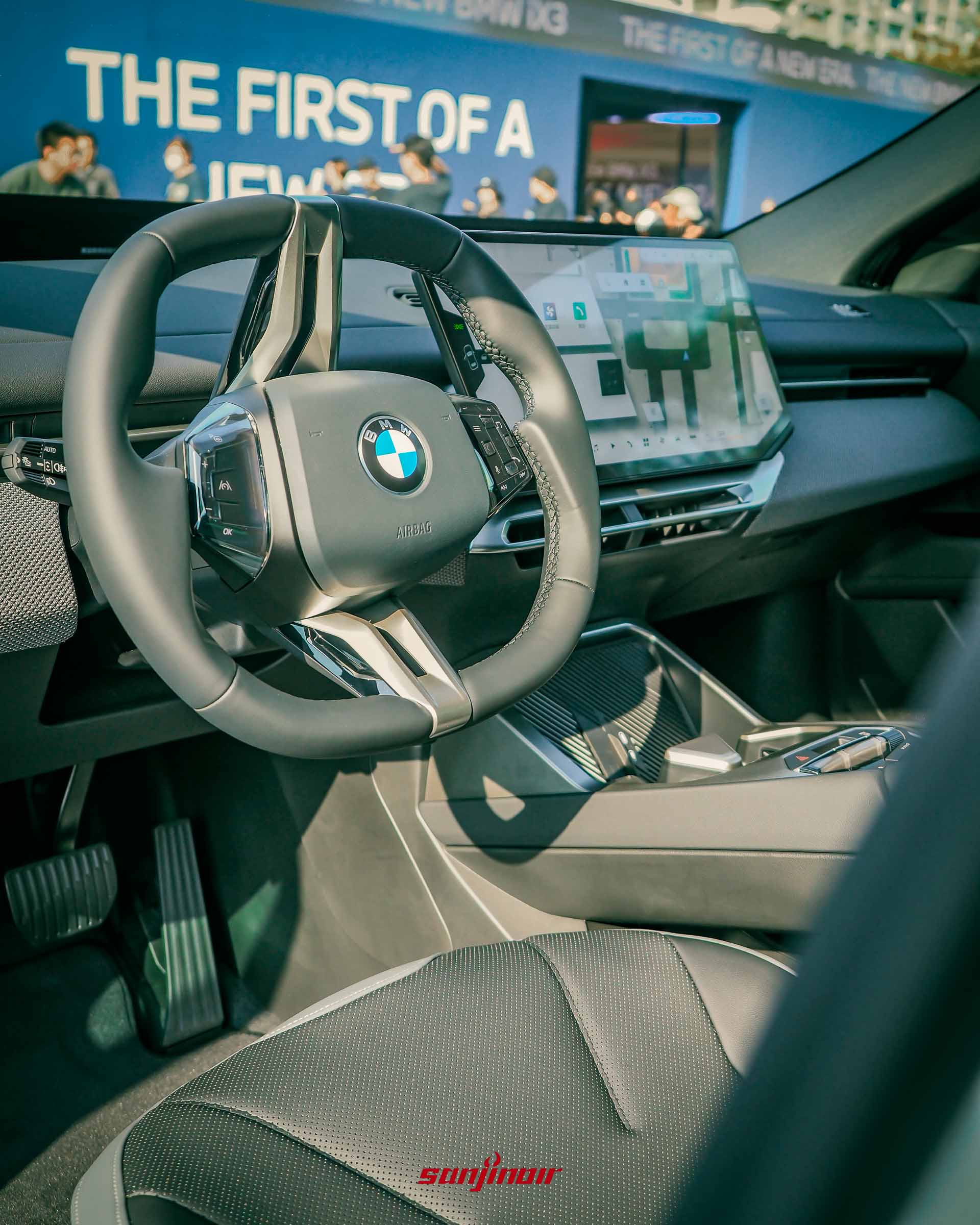 BMW iX3方向盤與中控螢幕特寫，窗外隱約可見展場「THE FIRST OF A NEW ERA」的標語。