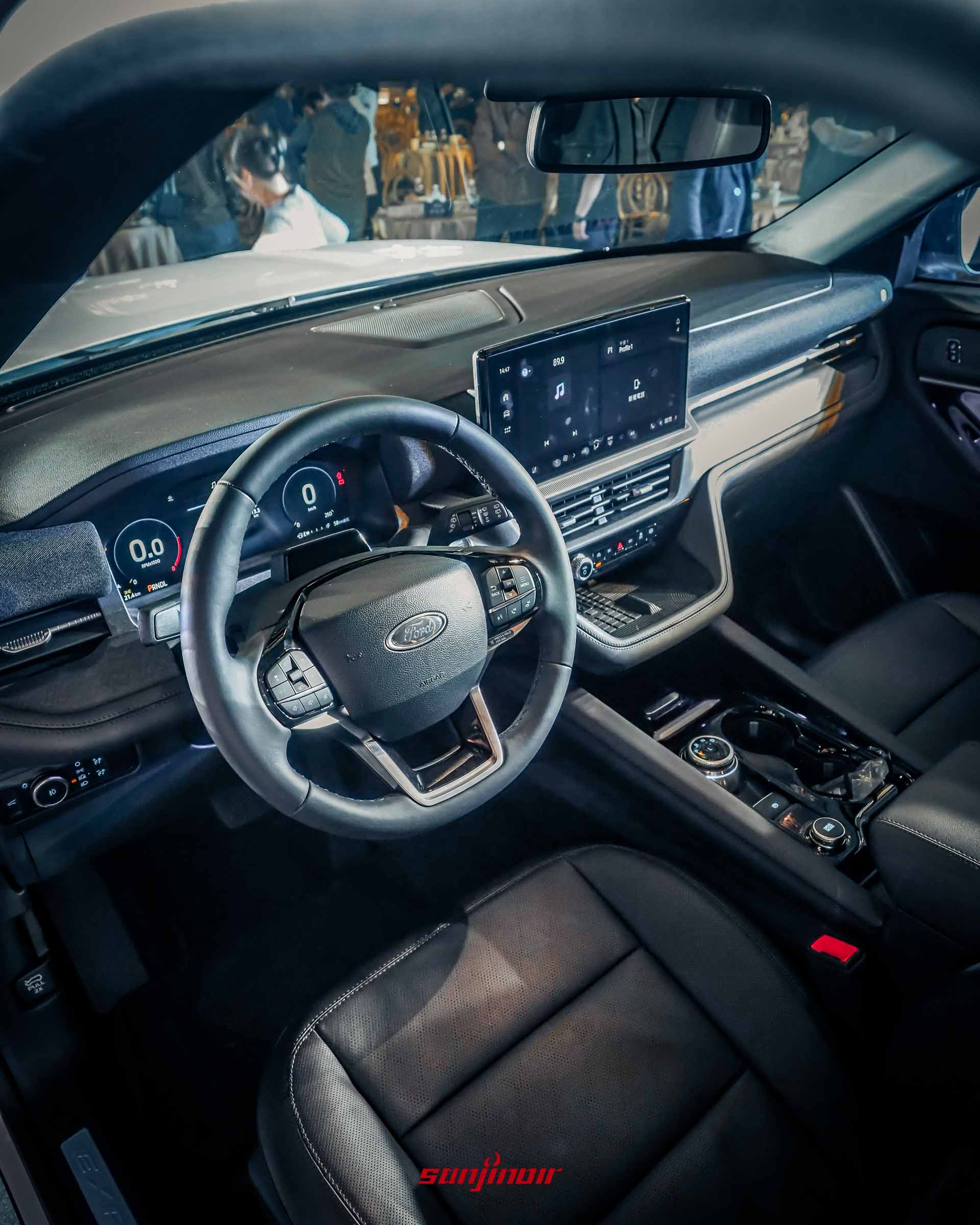 Ford Explorer車內駕駛艙俯視全景。黑色皮革方向盤位於前景，上有Ford Logo。後方是全數位儀表板和懸浮式中控大螢幕，顯示多媒體介面。中央扶手有旋鈕式換擋和杯架。黑色皮革座椅和車門飾板細節清晰。