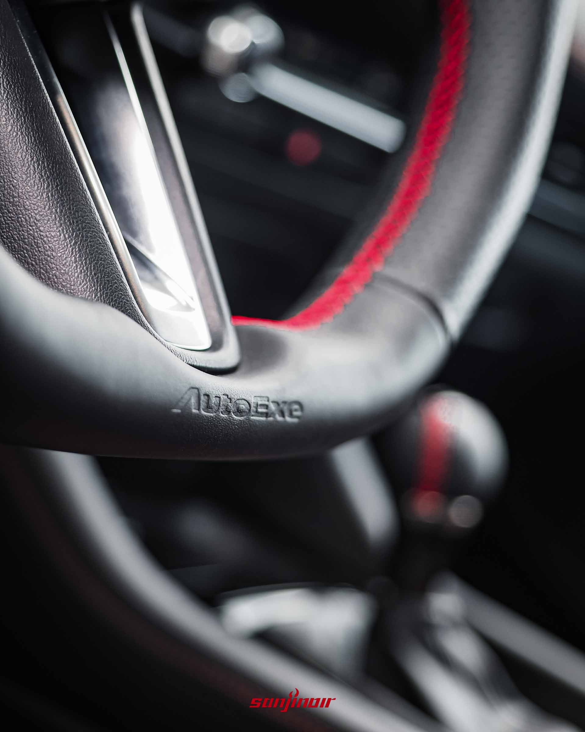 黑色AutoExe運動化平底方向盤底部刻有AutoExe標誌，背景有紅色縫線的球型排檔頭。