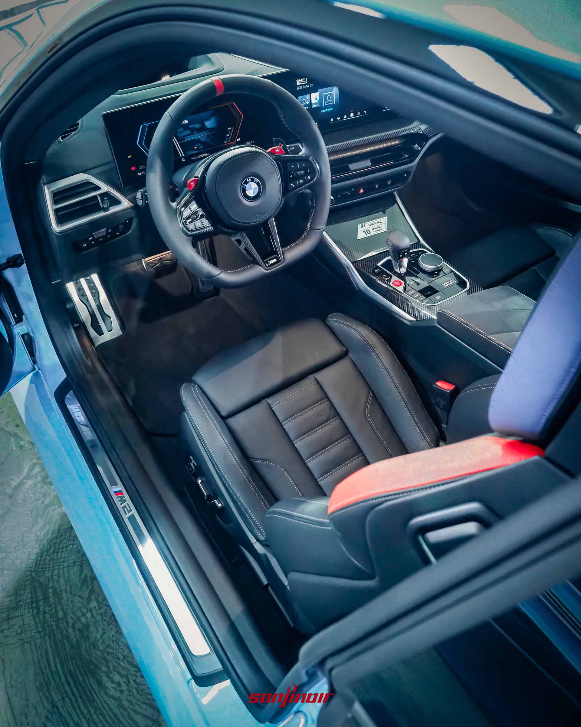 由車門外由上往下拍攝的BMW M2駕駛座艙視角照，展示多功能方向盤、雙螢幕中控台與碳纖維中央鞍座。