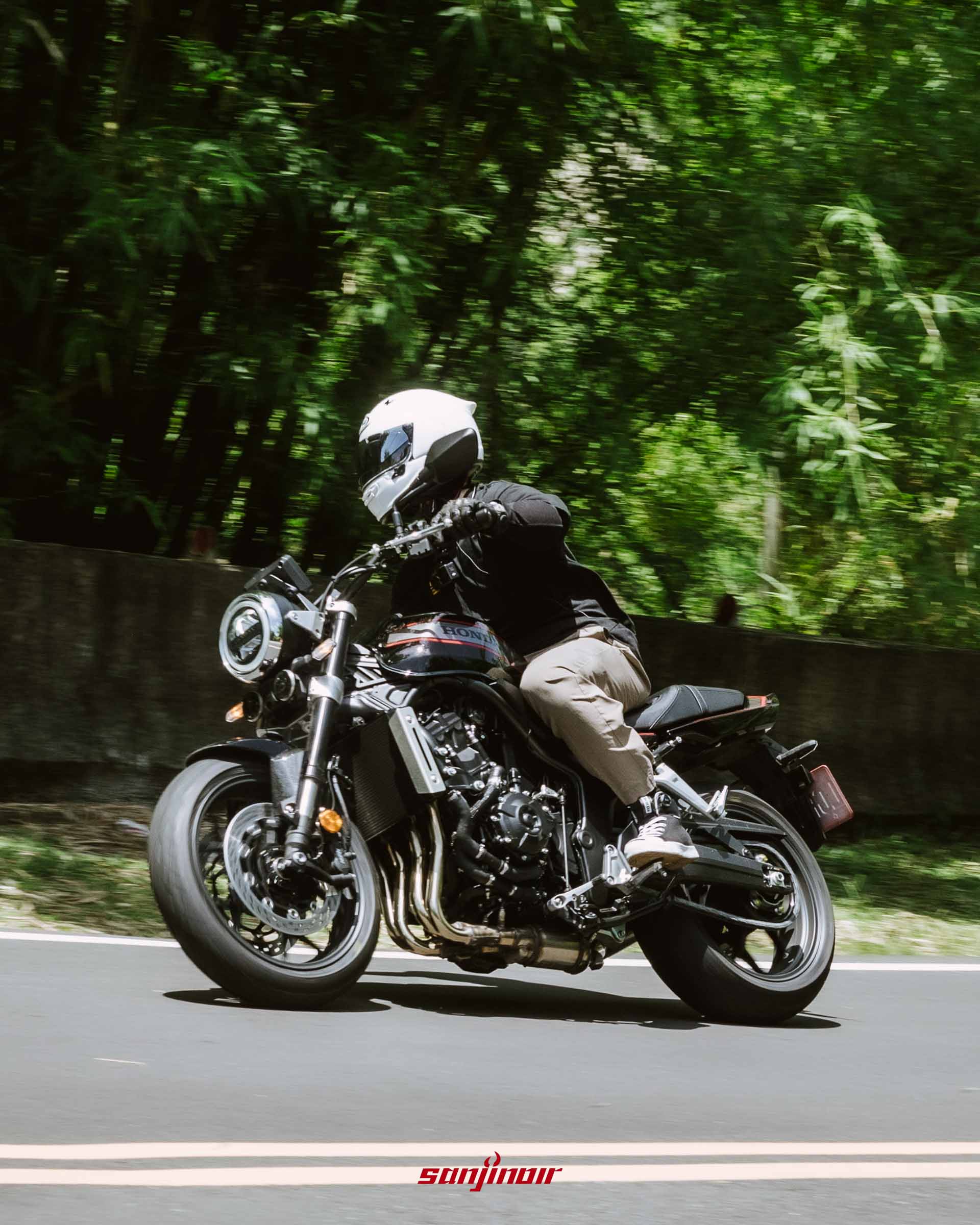 騎士佩戴白色安全帽騎乘黑色Honda CB1000F,在綠意盎然的山道中向右壓車過彎的動態追焦照片。
