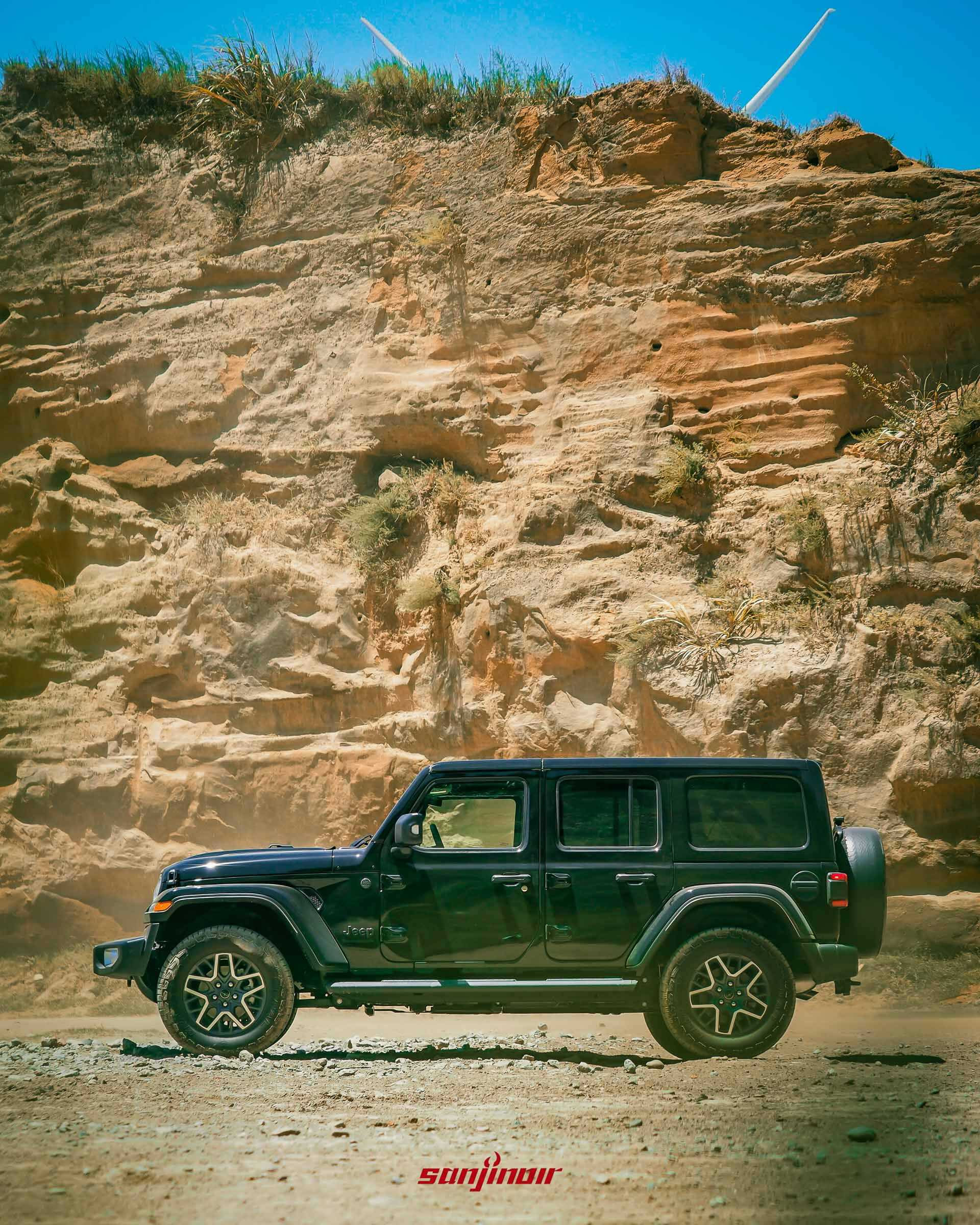 黑色 Jeep Wrangler Sahara 的側面全車照,停在陽光下的岩壁前,展示車身同色輪拱與車頂。