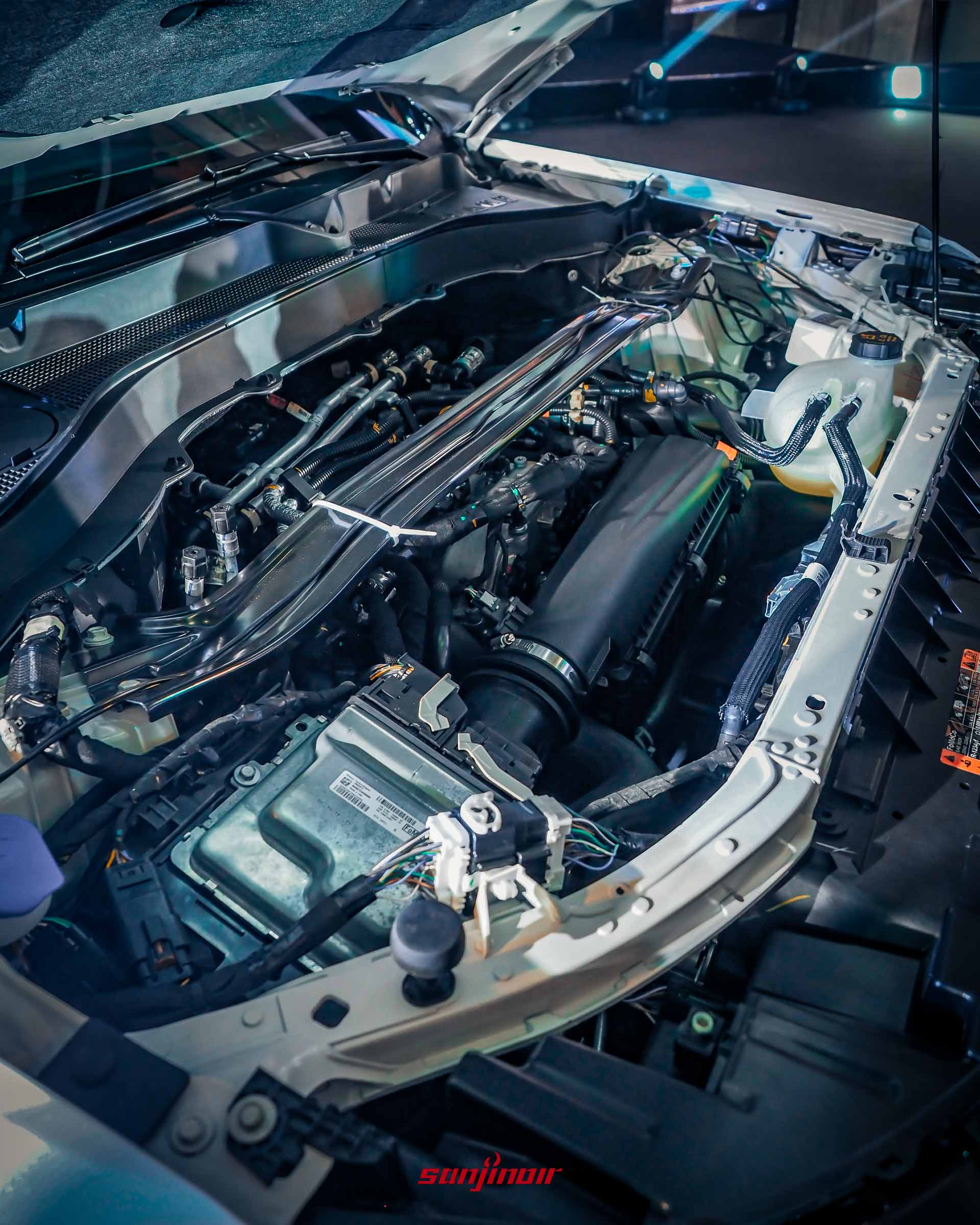 打開引擎蓋後的Ford Explorer引擎室近景特寫。可以看到2.3L EcoBoost引擎的頂部、各種管線、保險絲盒和紮帶固定細節。白色車身內部結構和隔音棉清晰可見。