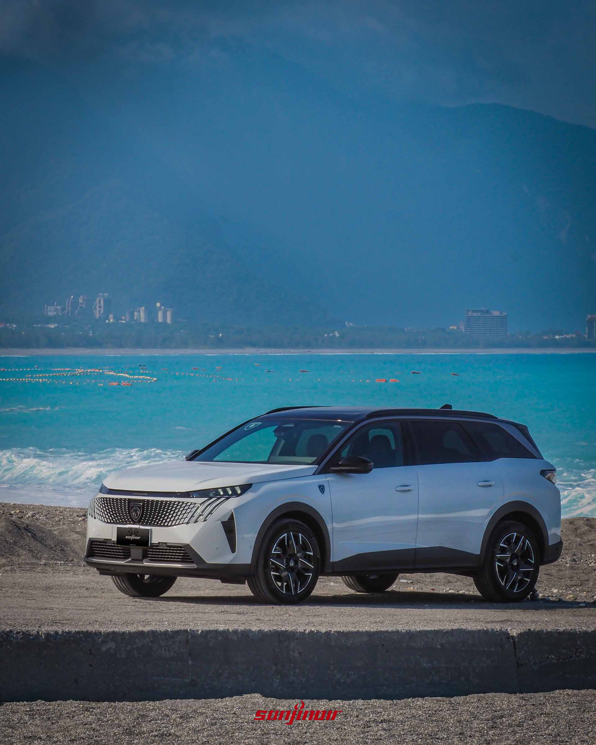 白色PEUGEOT 5008停放在海岸邊防波堤旁,背景是清澈湛藍的太平洋與遠方山脈及建築。