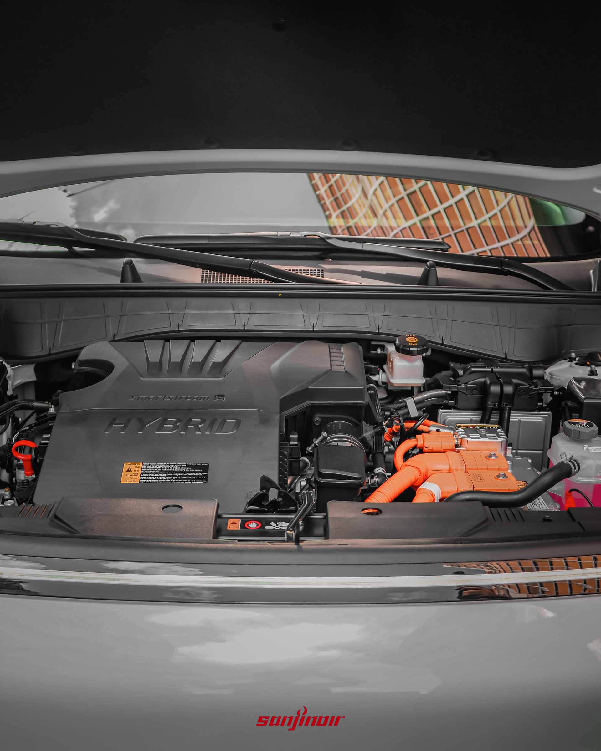 Kona Hybrid掀起引擎蓋後的引擎室特寫。畫面中央為帶有「HYBRID」與「Smartstream G」字樣的黑色引擎飾蓋,右側佈滿醒目的橘色高壓電線組、電瓶與各類動力控制模組。