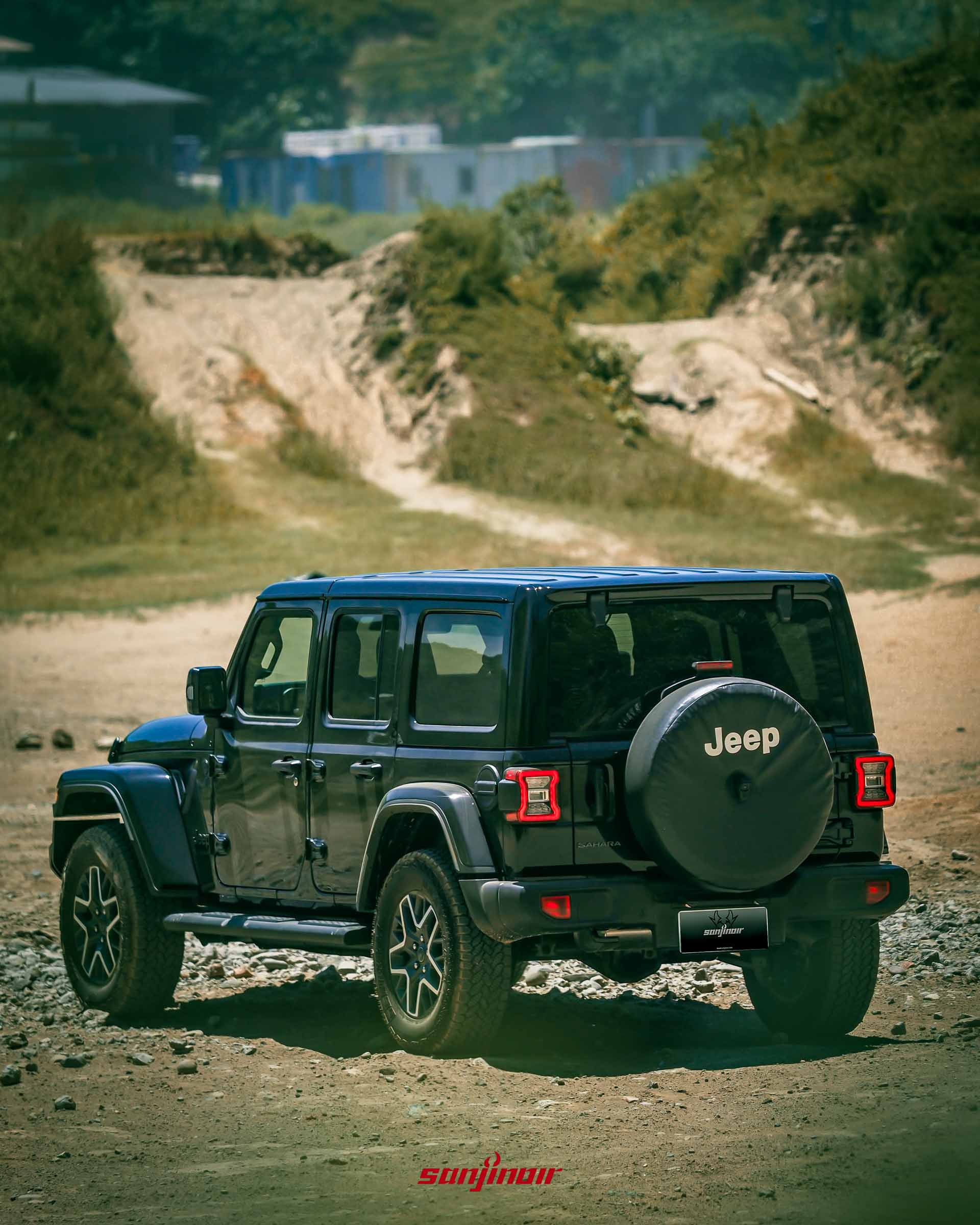 黑色 Jeep Wrangler Sahara 的右後方視角,停在泥土路面上,背景是綠色山坡,背著有 Jeep 字樣的備胎套。