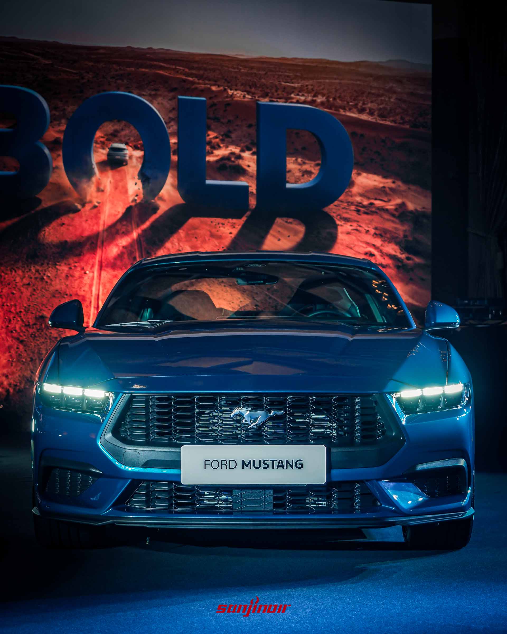 發表會舞台上藍色Ford Mustang的正臉特寫。LED頭燈亮起，黑色水箱護罩上有銀色野馬標誌。車牌顯示「FORD MUSTANG」。背景大螢幕顯示沙漠中一個巨大的「BOLD」字樣，光影投射在車身上。