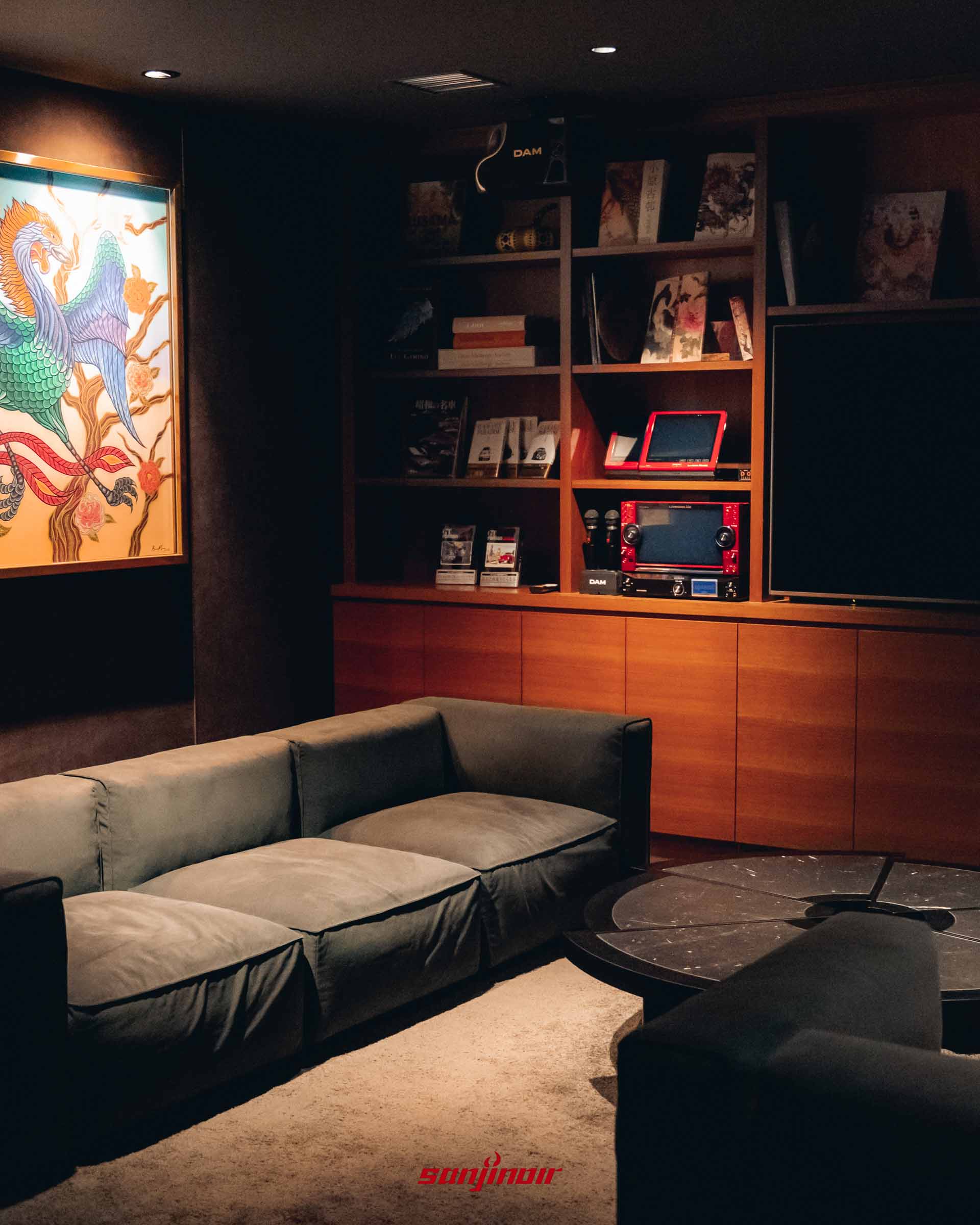 燈光昏暗的私人包廂內，設有深色絨面沙發、卡拉OK設備、書架以及牆上掛著鳳凰圖案的藝術畫作。