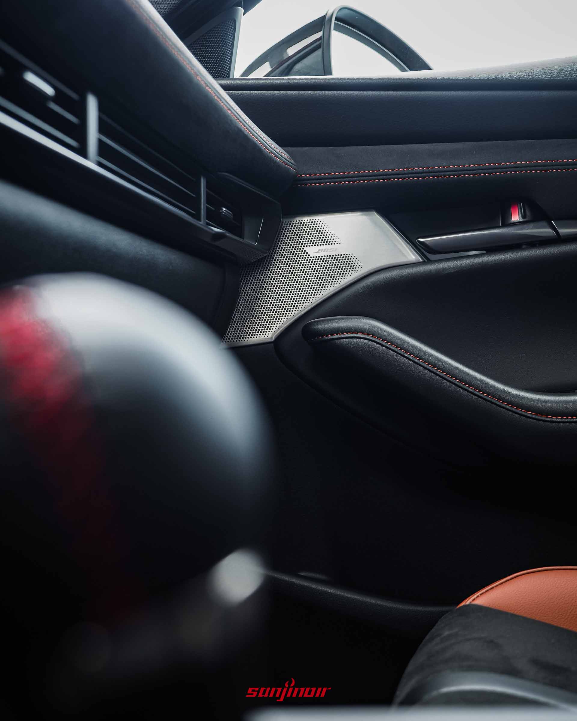 駕駛側車門特寫，有BOSE音響揚聲器護網，黑色皮革有紅色縫線，前景有模糊的排檔頭。