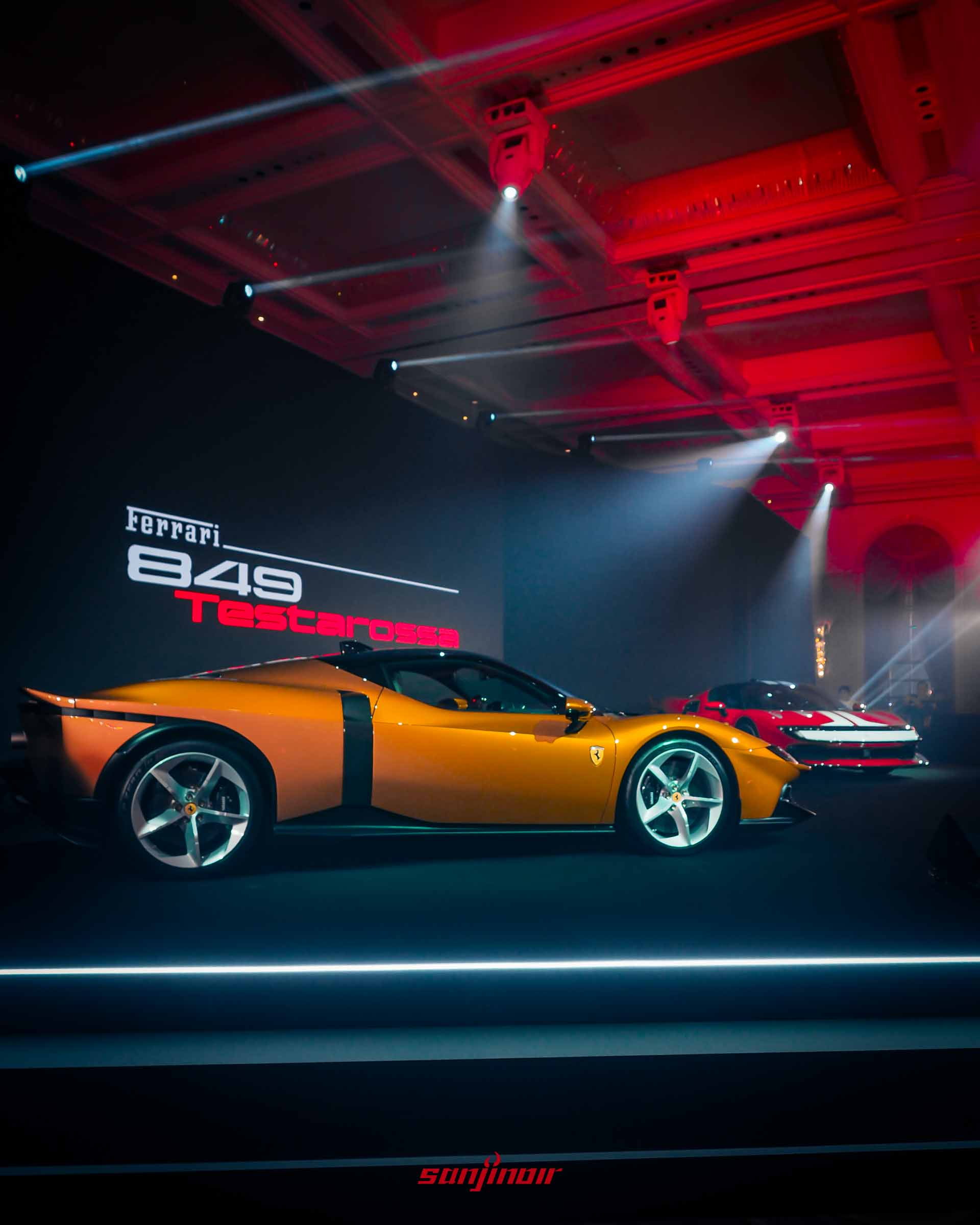 2026 Ferrari 849 Testarossa 橙色 Berlinetta 在發表會舞台上，燈光聚焦。車款側視角，背後的巨幅螢幕顯示著「Ferrari 849 Testarossa」字樣。舞台上還有紅色的 849 Testarossa 展示中。