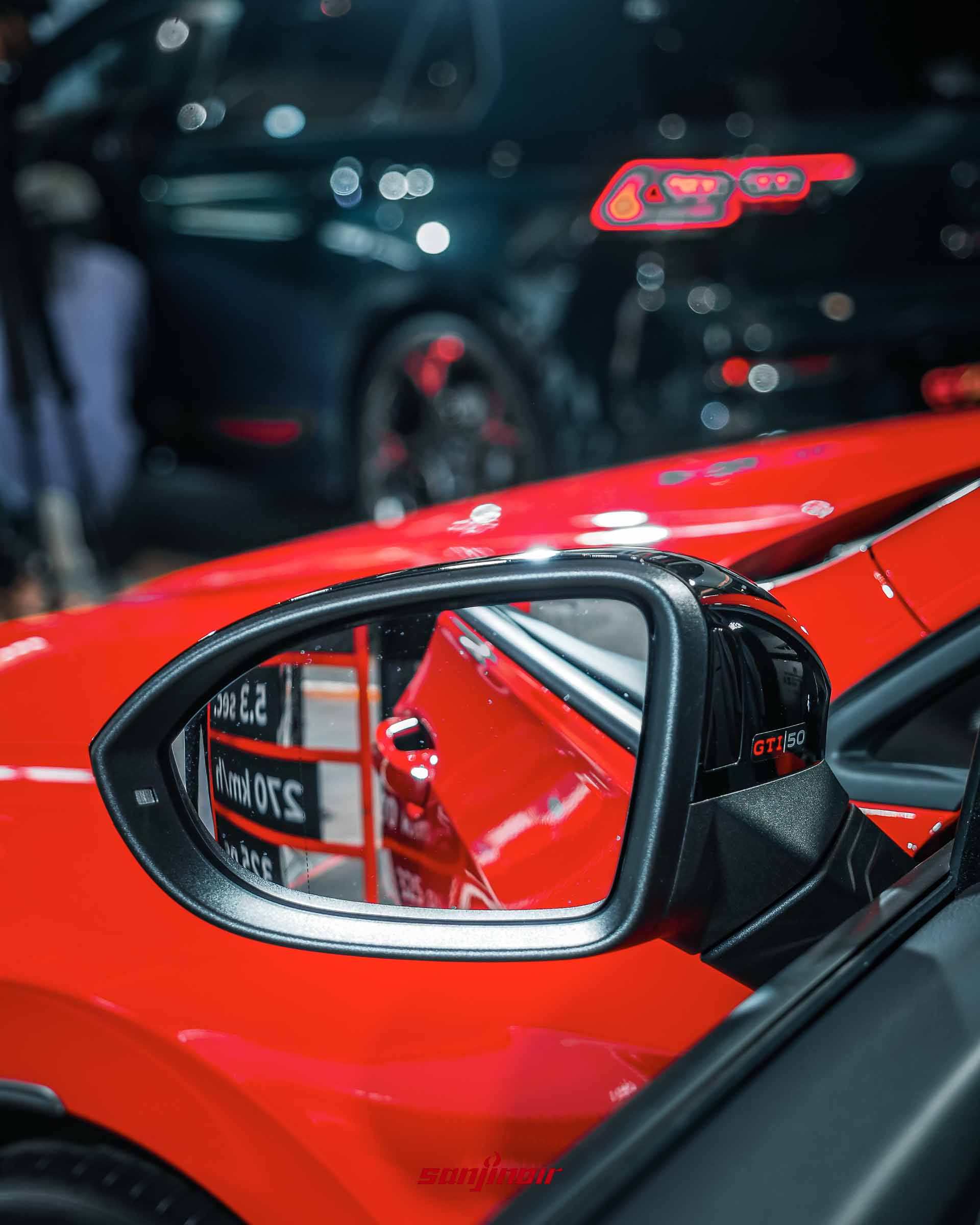 特寫Golf GTI EDITION 50黑化車外後視鏡，鏡殼上貼有GTI 50專屬徽飾，鏡面反射出現場紅黑配色的性能數據立牌。