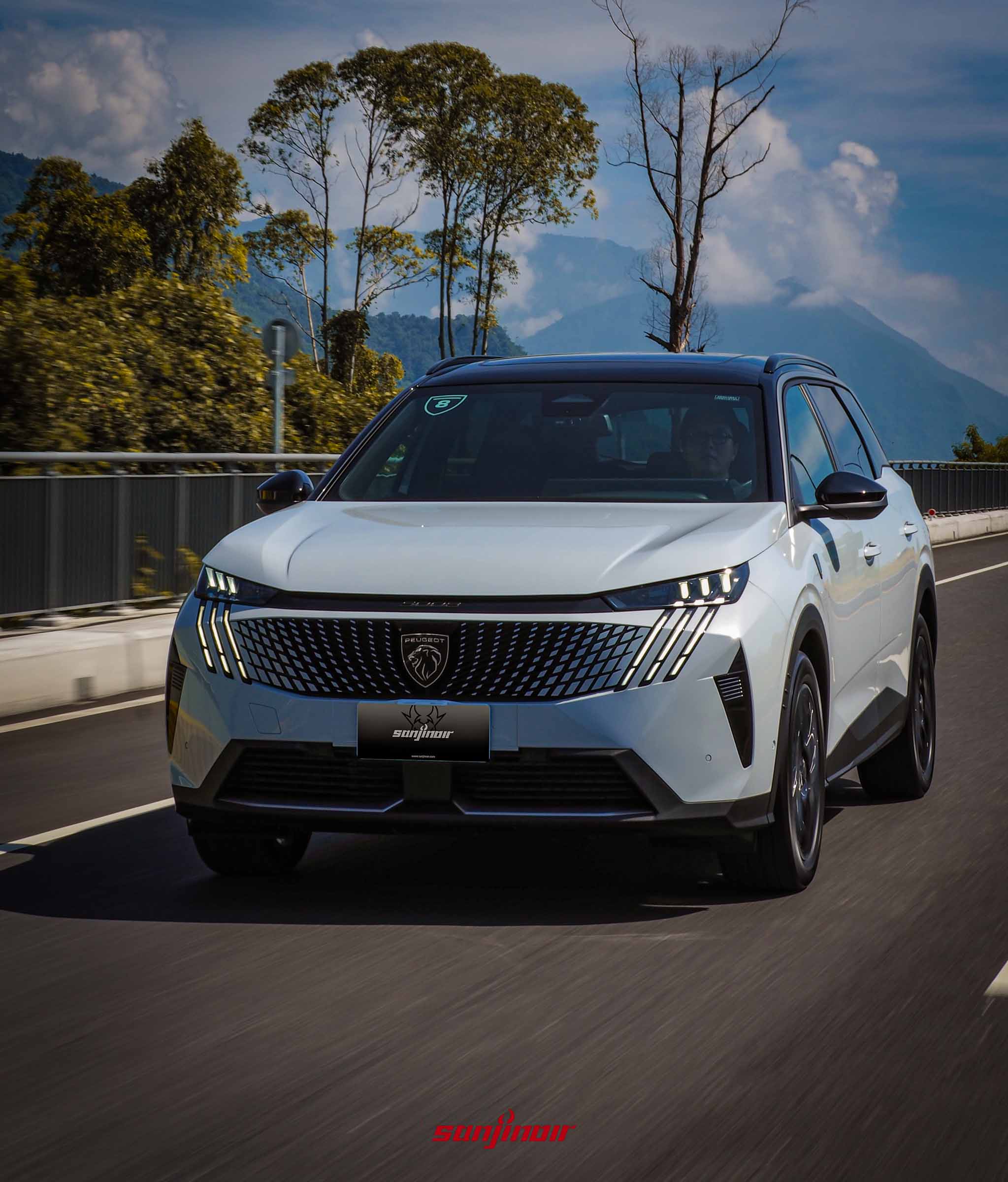白色PEUGEOT 5008在橋梁道路上行駛的動態照,背景為藍天白雲與連綿的翠綠山脈。