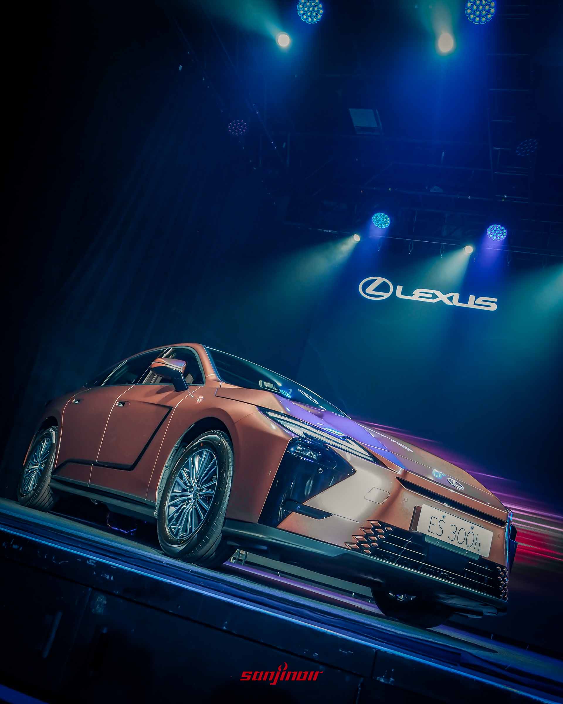 舞台上銅色Lexus ES300h低角度正面照，展示車頭造型與多輻式鋁圈設計，背景有Lexus品牌標誌。