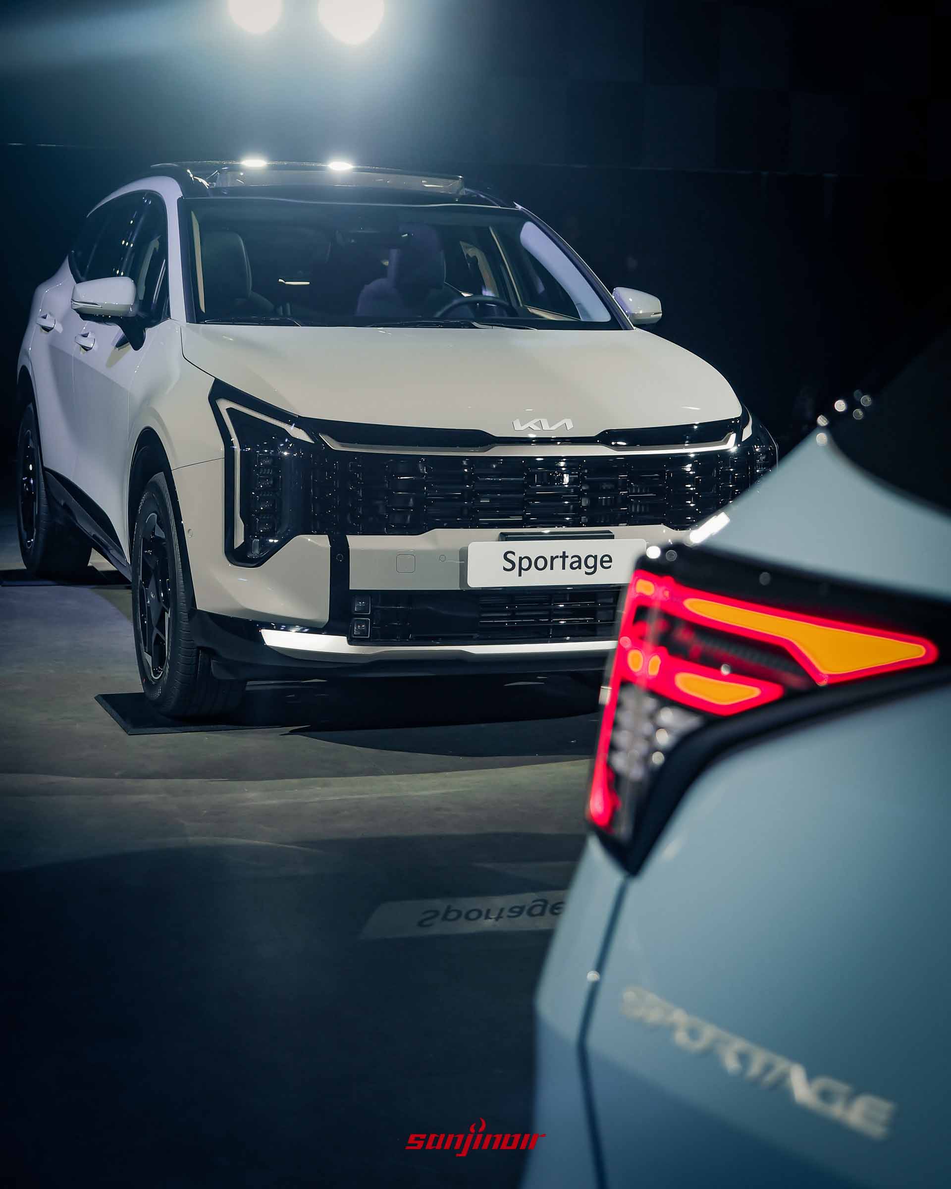 同台展示兩台Kia Sportage，前方為一台珍珠白車型的車頭，後方為一台晨霧藍車型的車尾，前景有白色車頭和日行燈光條。