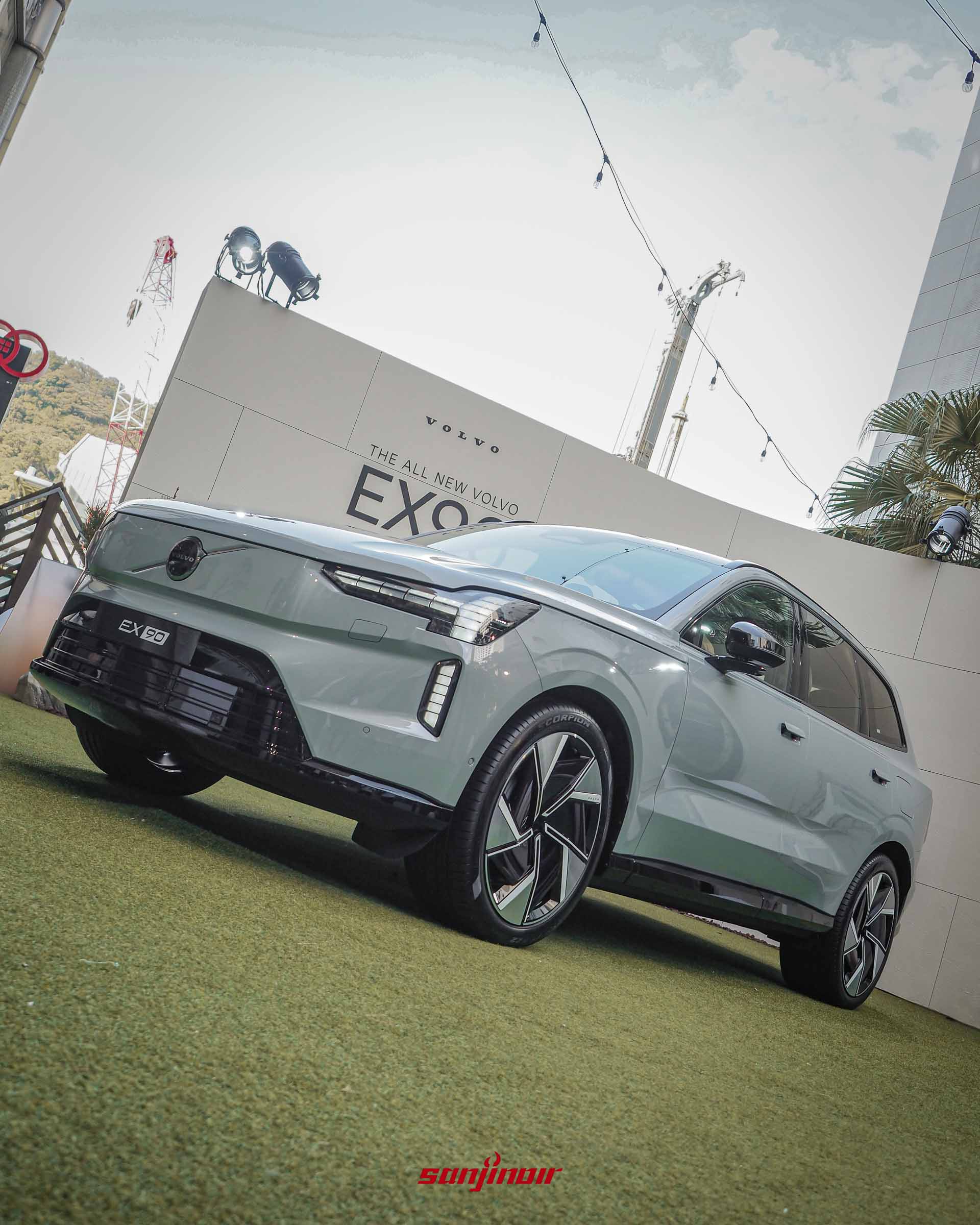 由低角度拍攝的灰色Volvo EX90,車頭朝向左前方,強調其休旅車高大挺拔的氣勢與俐落的車身折線,背景為發表會活動場地及燈光設備。