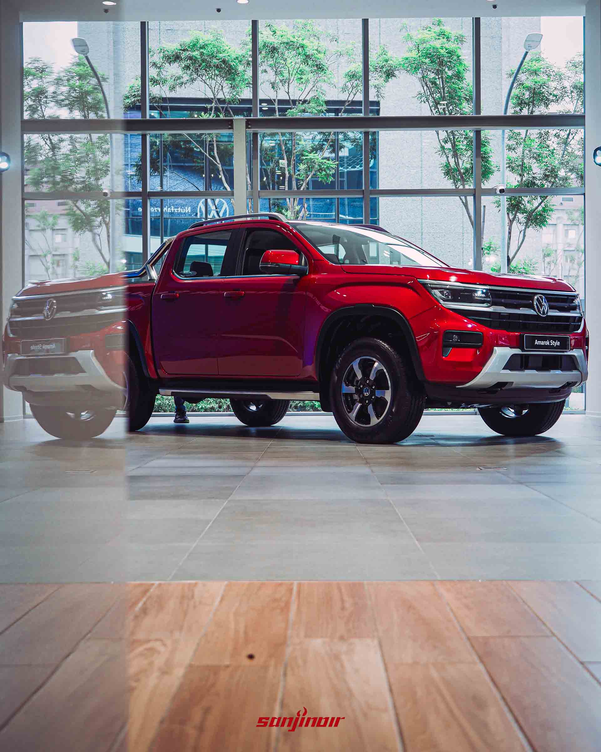 紅色Volkswagen Amarok Style停放於明亮展間的側視圖，前景帶有木質地板與玻璃反光，背景為落地窗外景。