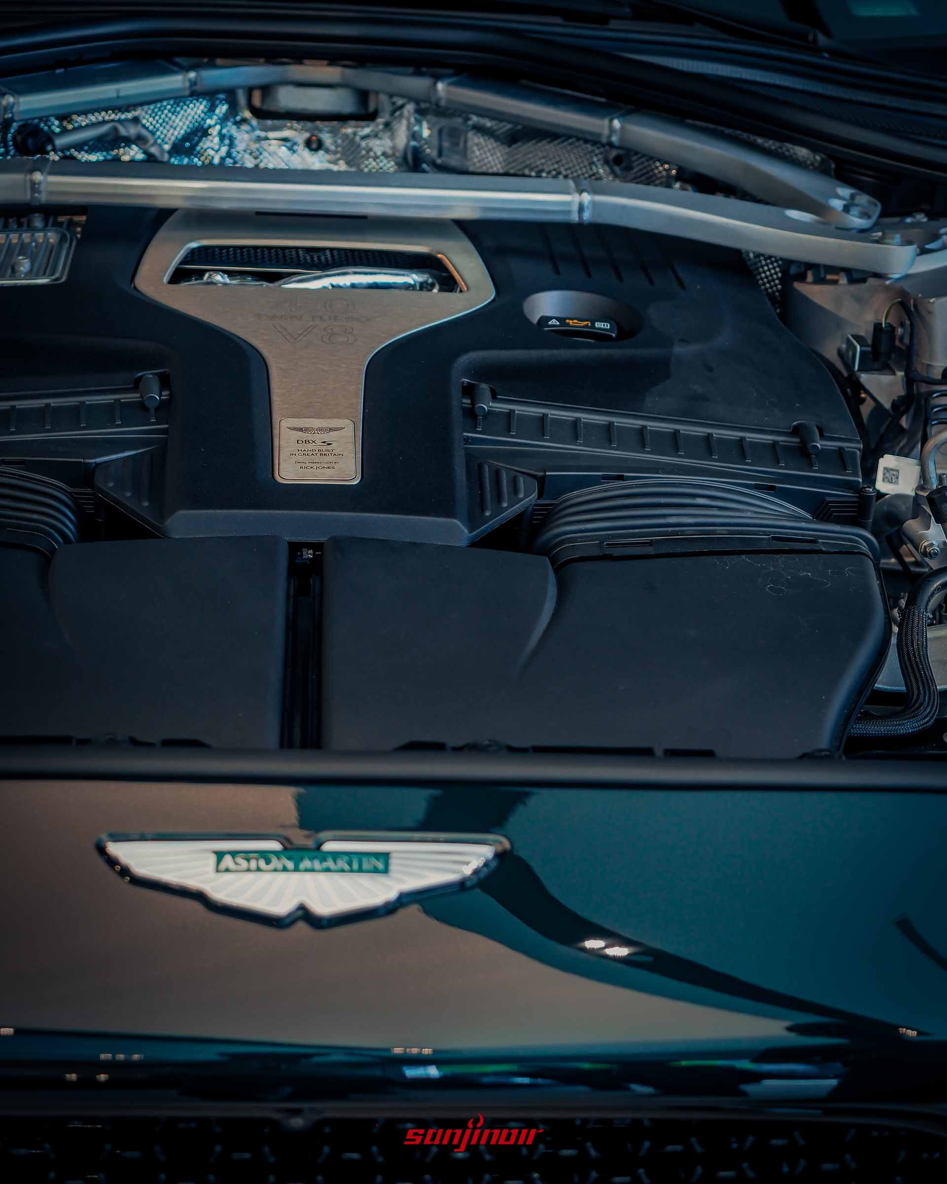 Aston Martin DBX S引擎室特寫，展示V8雙渦輪增壓引擎護蓋、碳纖維拉桿、Aston Martin飛翼廠徽以及刻有工程師簽名的英國手工組裝銘牌。