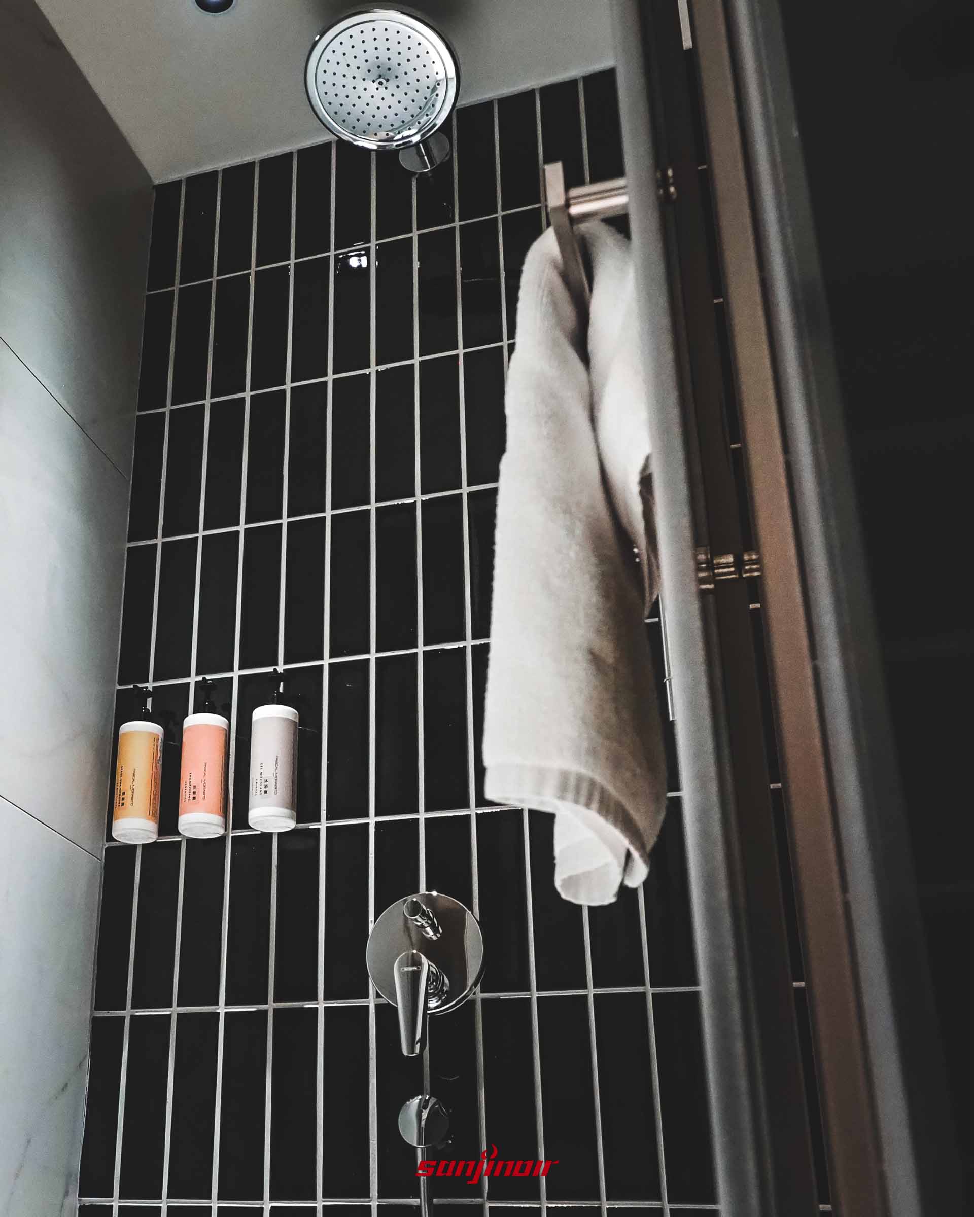 淋浴間內部,黑色磁磚牆面上裝有金屬花灑及三罐洗沐備品,玻璃門把上掛著毛巾。
