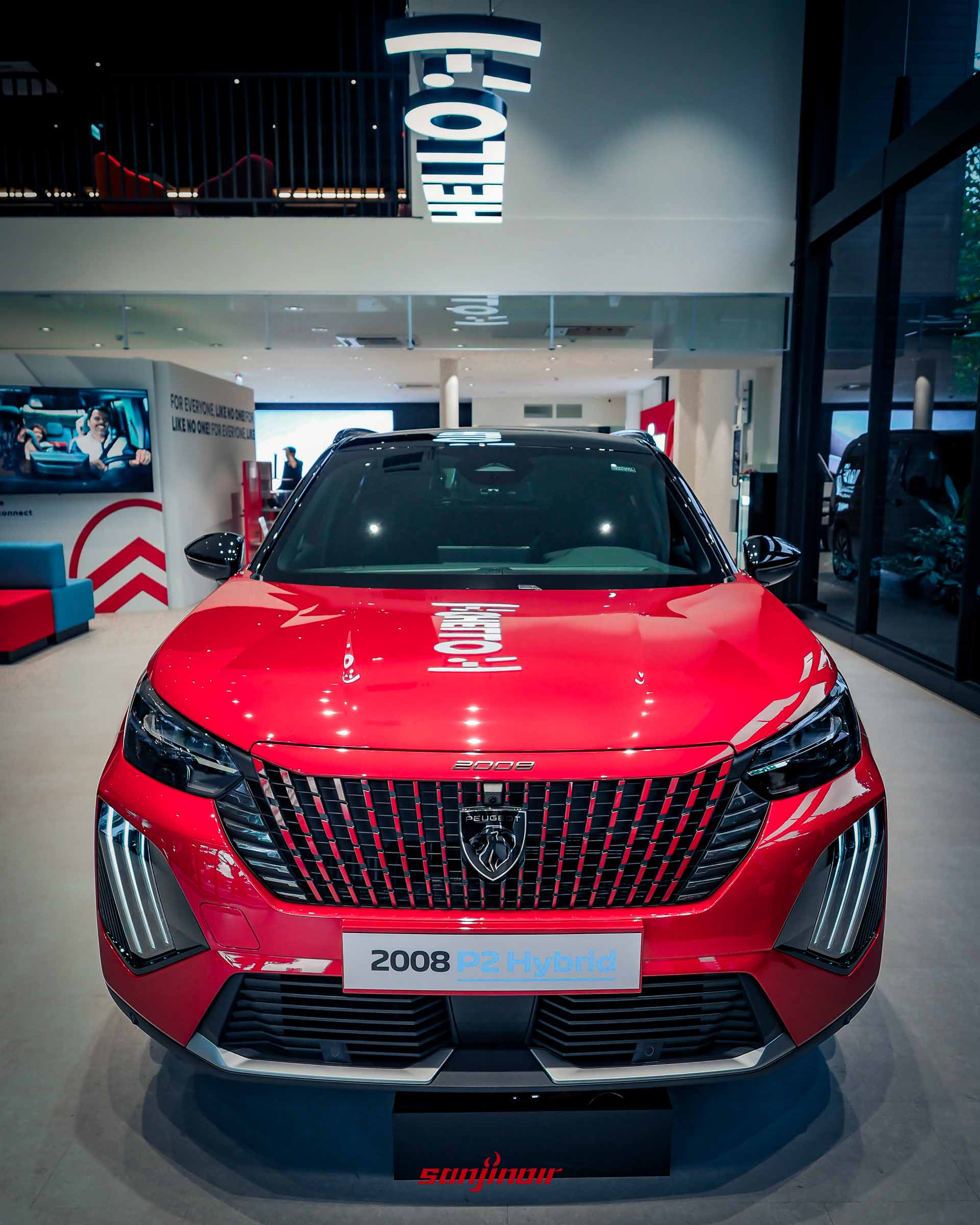 一輛亮紅色 PEUGEOT 2008 P2 Hybrid 小型 SUV 的車頭正視圖，位於一個汽車展廳內。車輛具有新款無邊框格柵設計，中央是新的「Peugeot」獅子標誌，以及標誌性的垂直爪痕 LED 日行燈。