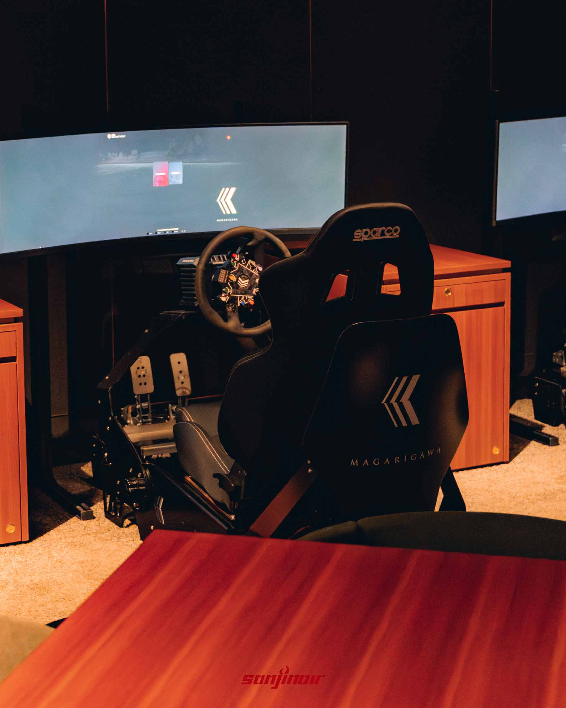 一套配備超寬曲面螢幕、方向盤與金屬踏板的賽車模擬器，黑色賽車椅背上繡有 MAGARIGAWA 標誌。