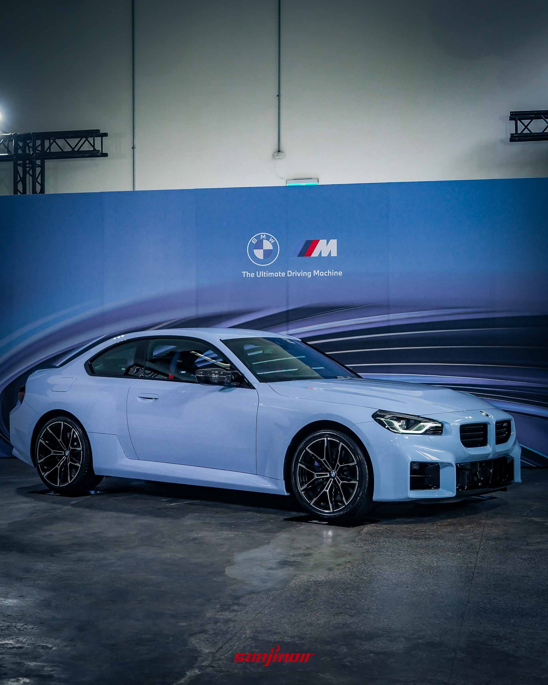 淺藍色BMW M2 M xDrive右前方45度角外觀照，停放於室內展演空間，背景有BMW與M品牌標誌背板。