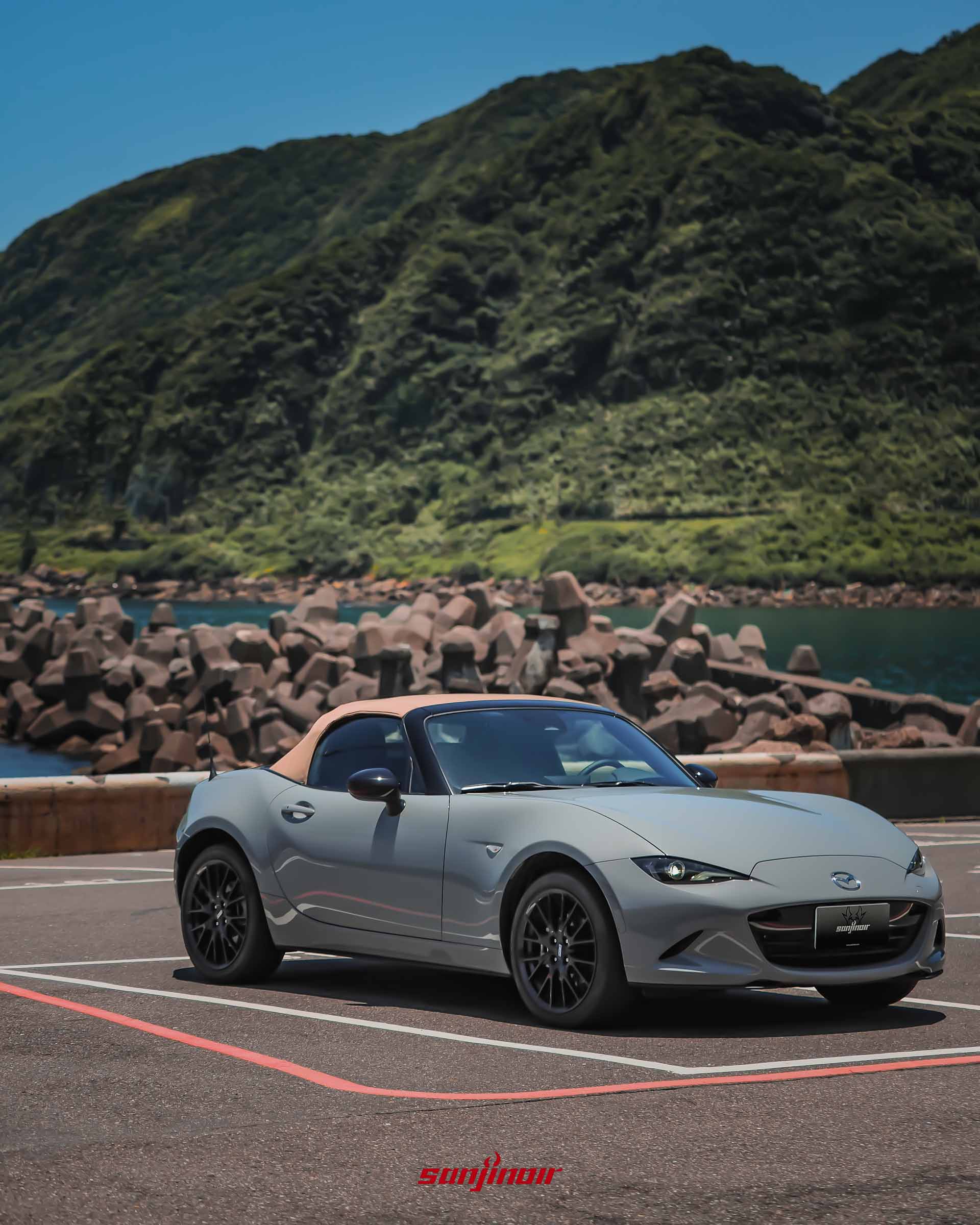 灰色MAZDA MX-5 RS關篷狀態,配備棕色軟式頂篷,停放於消波塊與翠綠山峰交織的海岸旁。