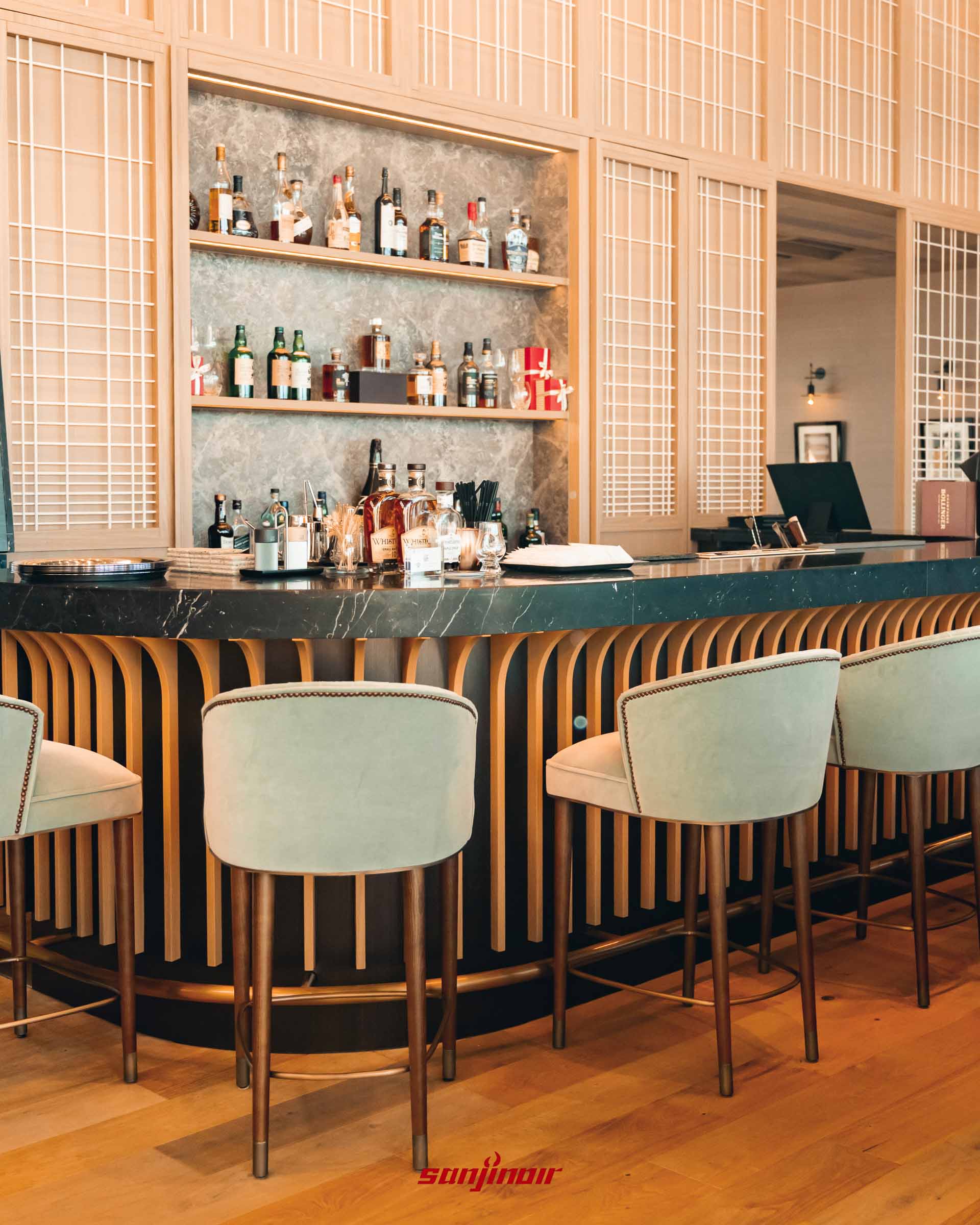 高質感的吧檯區域，設有黑色大理石檯面、木質格柵裝飾與淺綠色絨布高腳椅，後方層架擺滿各式烈酒。