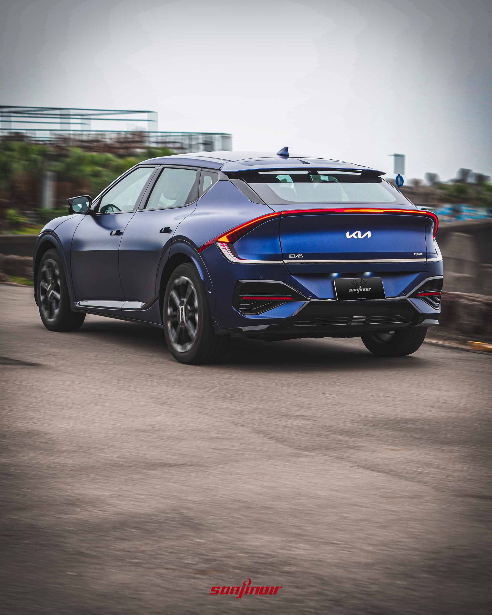 藍色啞光Kia EV6 GT-Line電動車在道路上行駛的動態追焦照片，從右後方視角展示亮起的橫貫式尾燈與行進姿態。