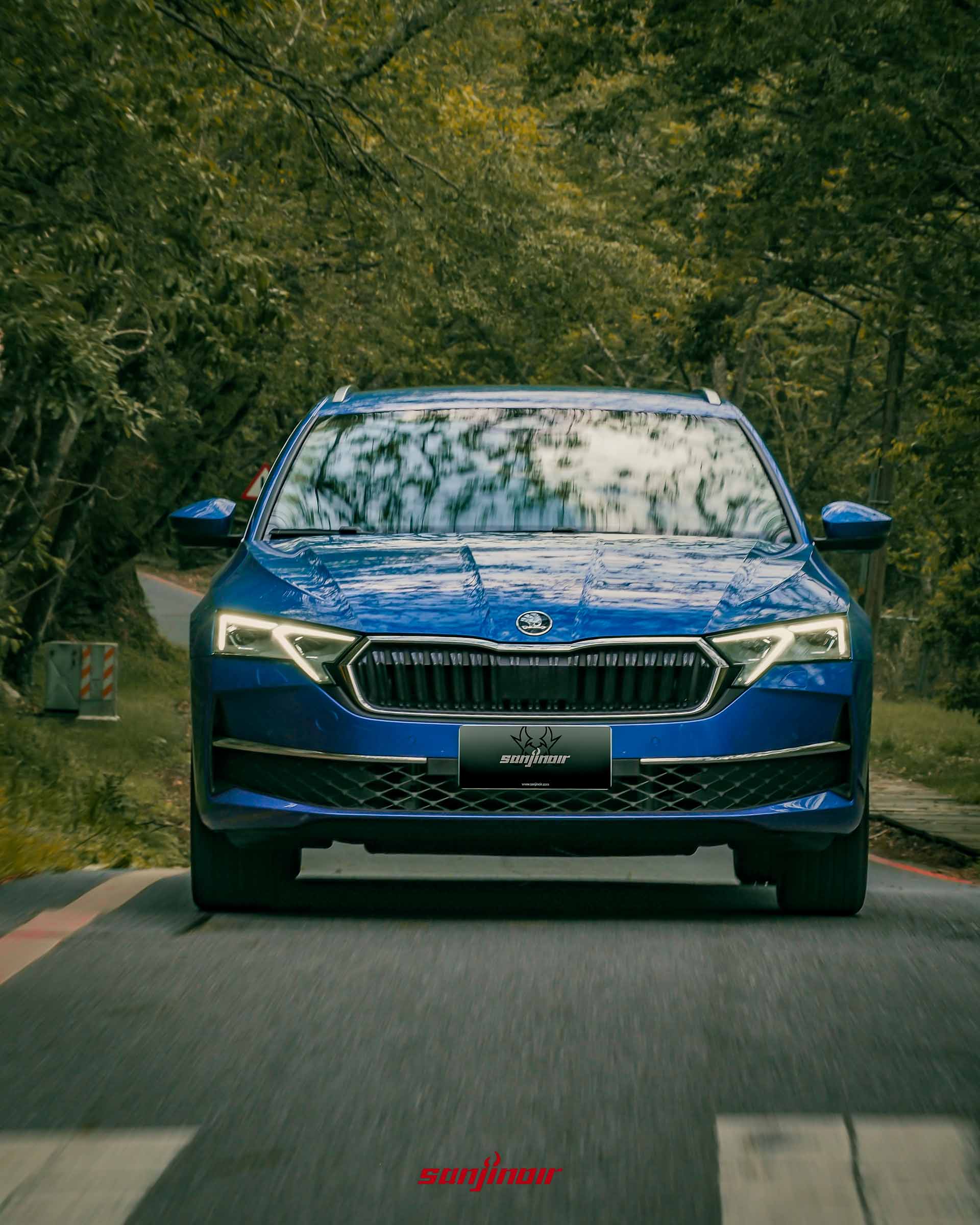藍色 Škoda Octavia Combi 2.0 TSI 4×4 行駛在山路上的正面特寫照片，頭燈亮起，背景是模糊的樹林。