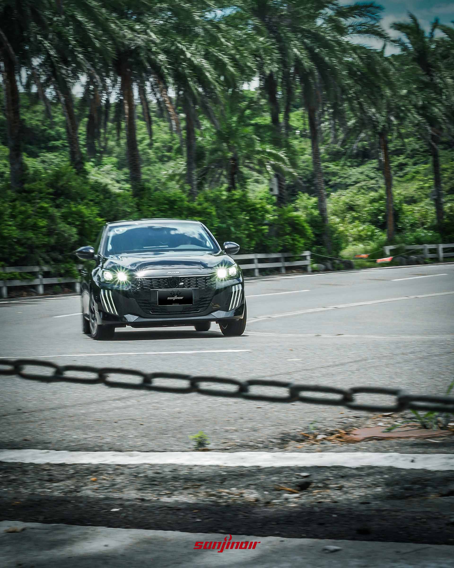 黑色PEUGEOT 208 Hybrid行駛於充滿綠意的蜿蜒山路正面動態追焦照。