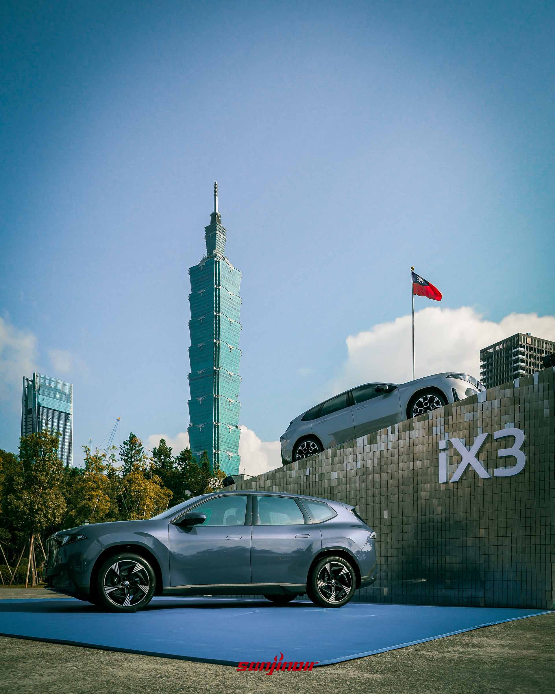 兩輛BMW iX3於戶外展出，一輛深藍色在平地，一輛銀色在階梯狀展台，背景矗立著地標台北101大樓。