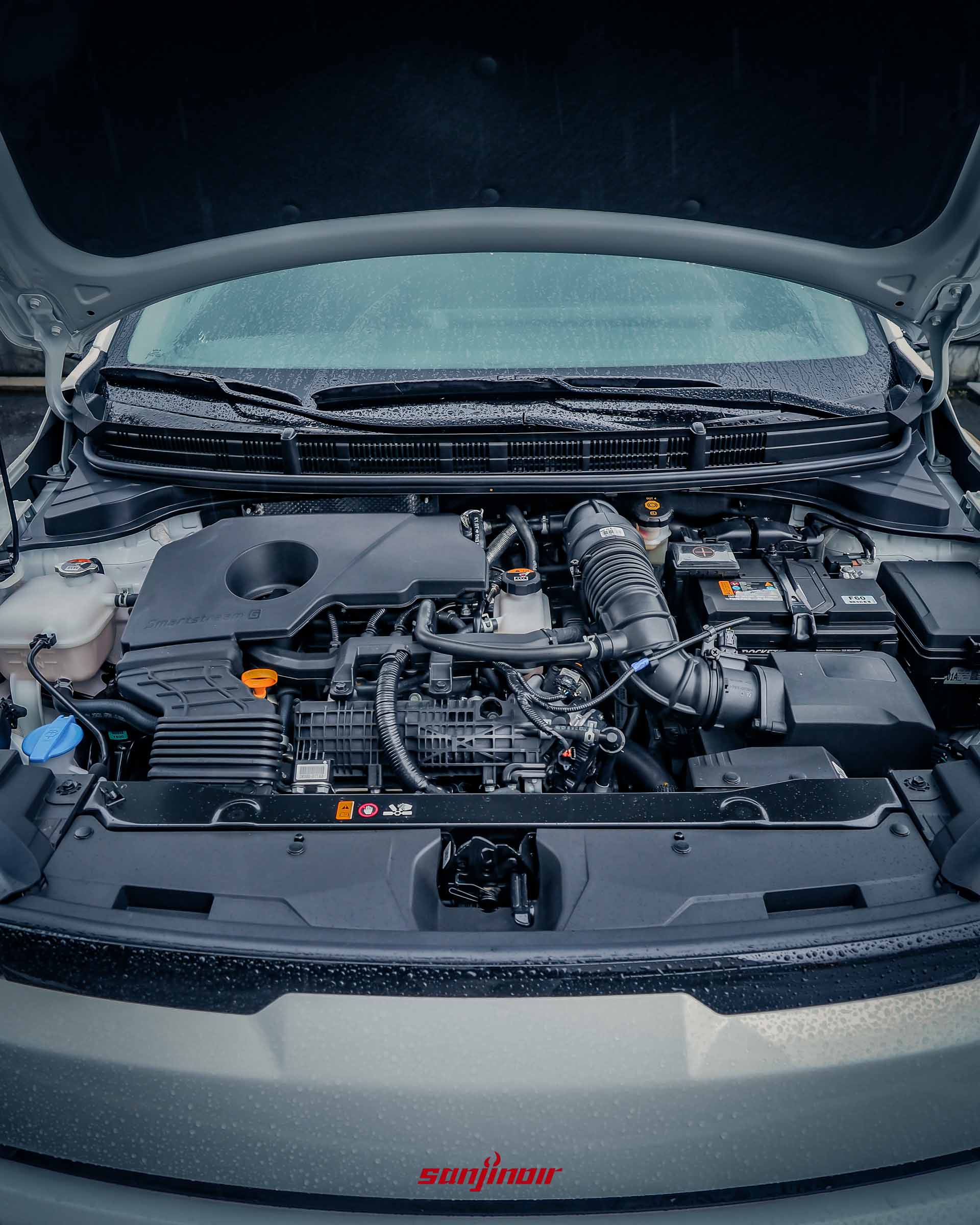 開啟引擎蓋後正面俯視 2026 Kia Stonic 的引擎室,展示帶有 Smartstream G 標誌的 1.0L 三缸渦輪增壓引擎本體與 48V MHEV 智慧輕油電系統相關部件,車身帶有雨水。