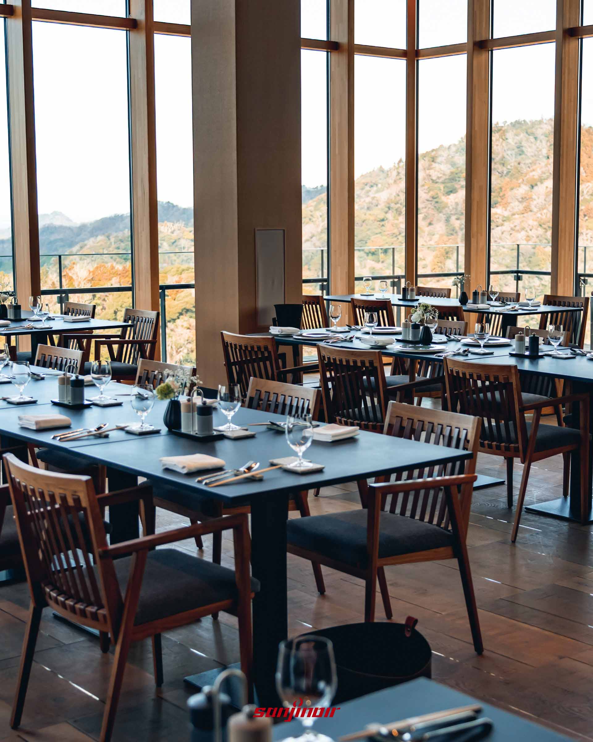 餐廳內部視角，深色木質餐桌上擺放著整齊的餐具與水杯，陽光透過大片落地窗灑落，窗外為蓊鬱的山林景色。