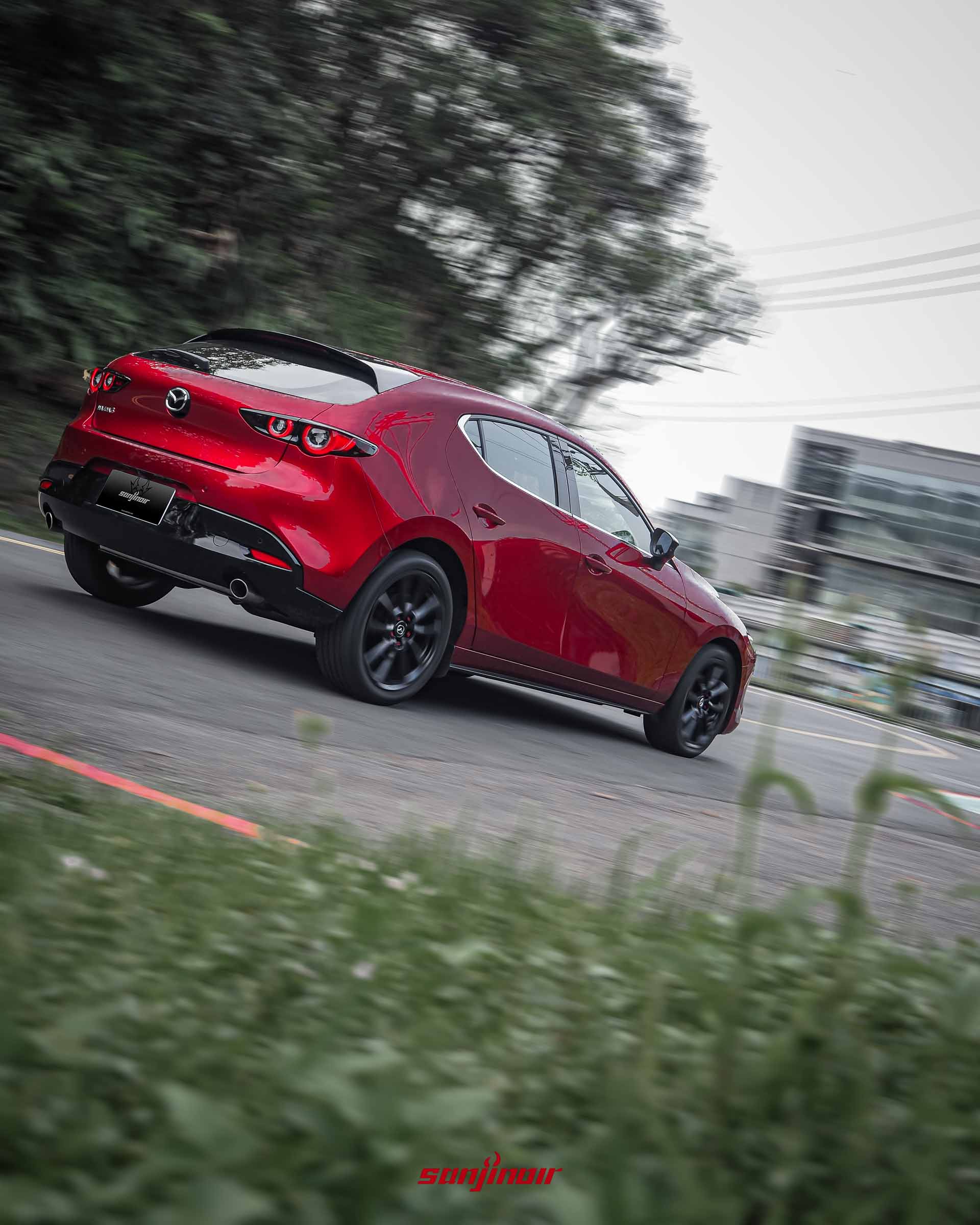 晶艷魂動紅MAZDA3 HOMURA在道路上行駛的動態追焦照，從右後方視角拍攝，車身線條流暢，背景帶有速度感的模糊效果。