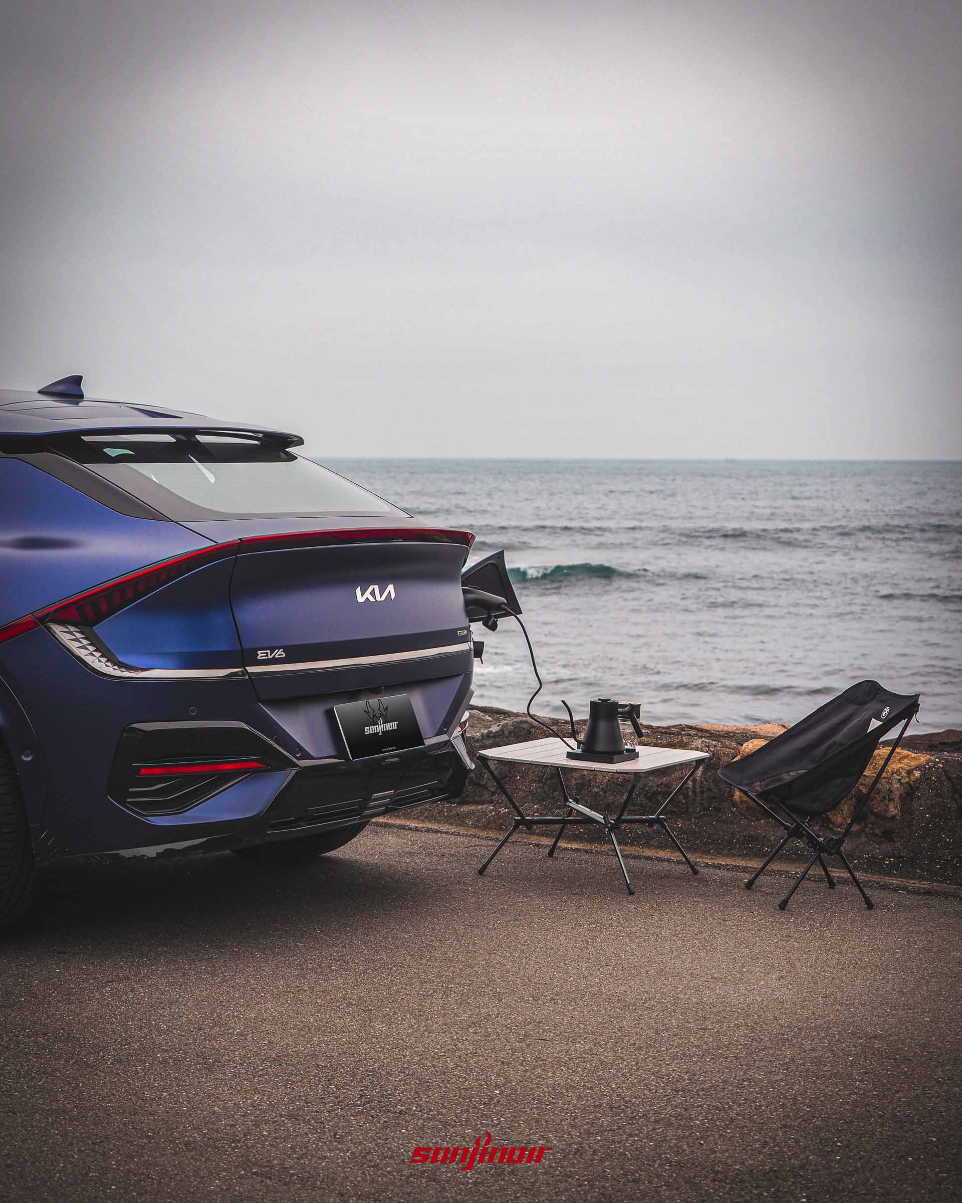 Kia EV6 GT-Line小改款停在海邊，車尾充電孔接出電纜供電給戶外桌上的電水壺，旁邊擺放露營椅，展示V2L供電功能。