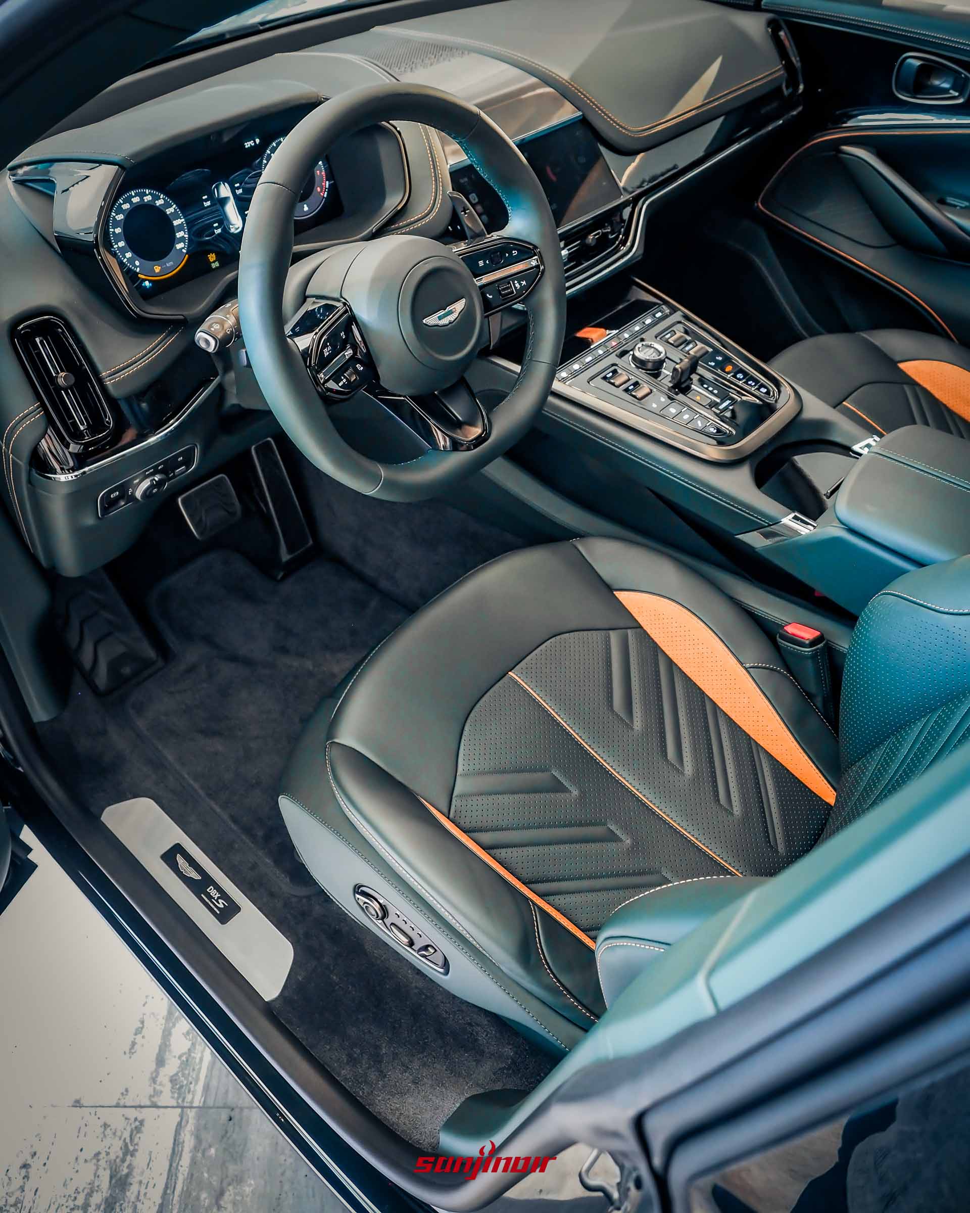 Aston Martin DBX S駕駛座艙全景，從車門視角看入，展示深色皮革搭配橘色縫線與點綴的跑車座椅、數位儀表板、跑車化方向盤與帶有DBX S字樣的金屬迎賓踏板。