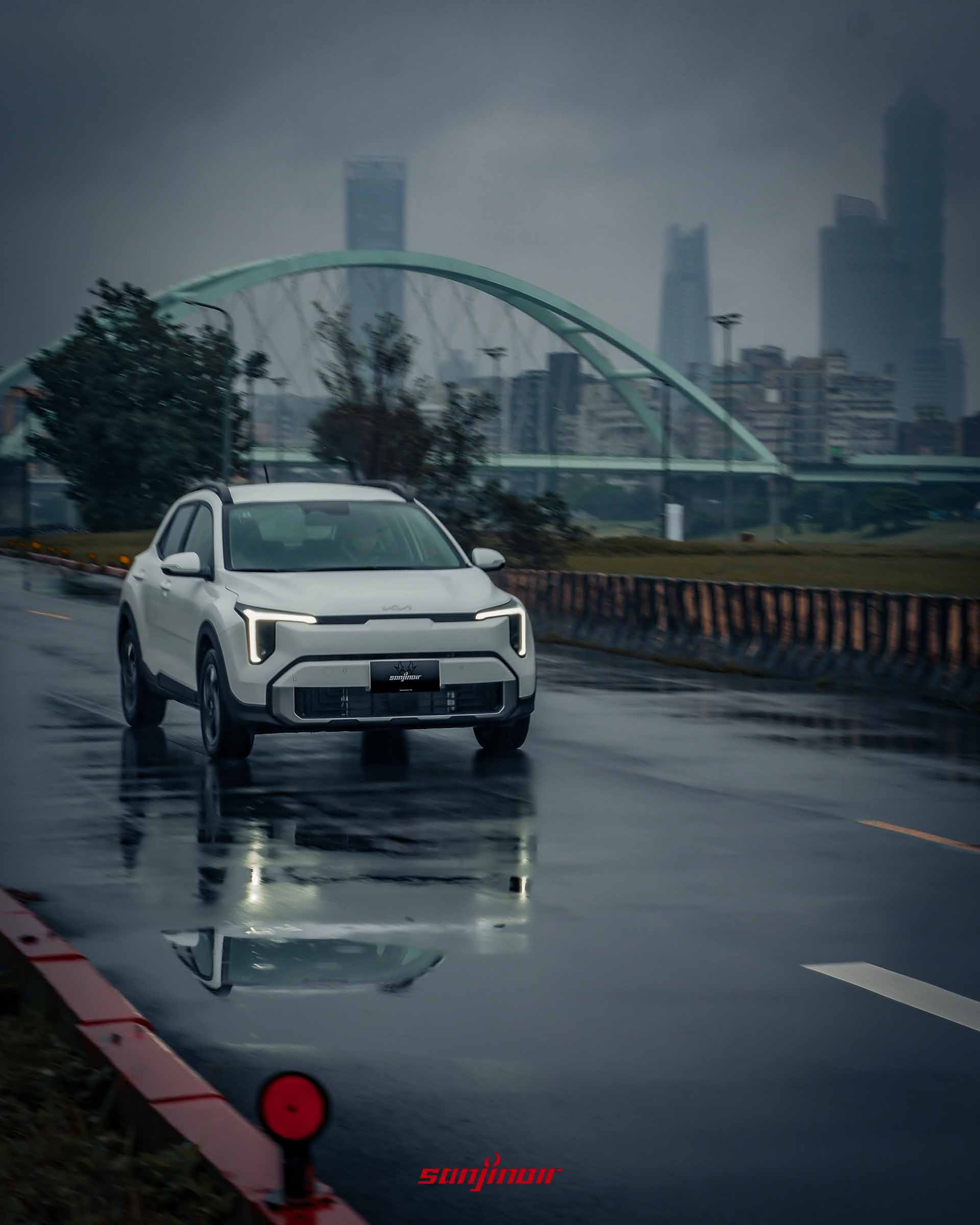 白色 2026 Kia Stonic 在陰雨天動態行駛於濕滑的道路上,路面有車輛倒影,背景為模糊的城市天際線與樹木,頭燈亮起。