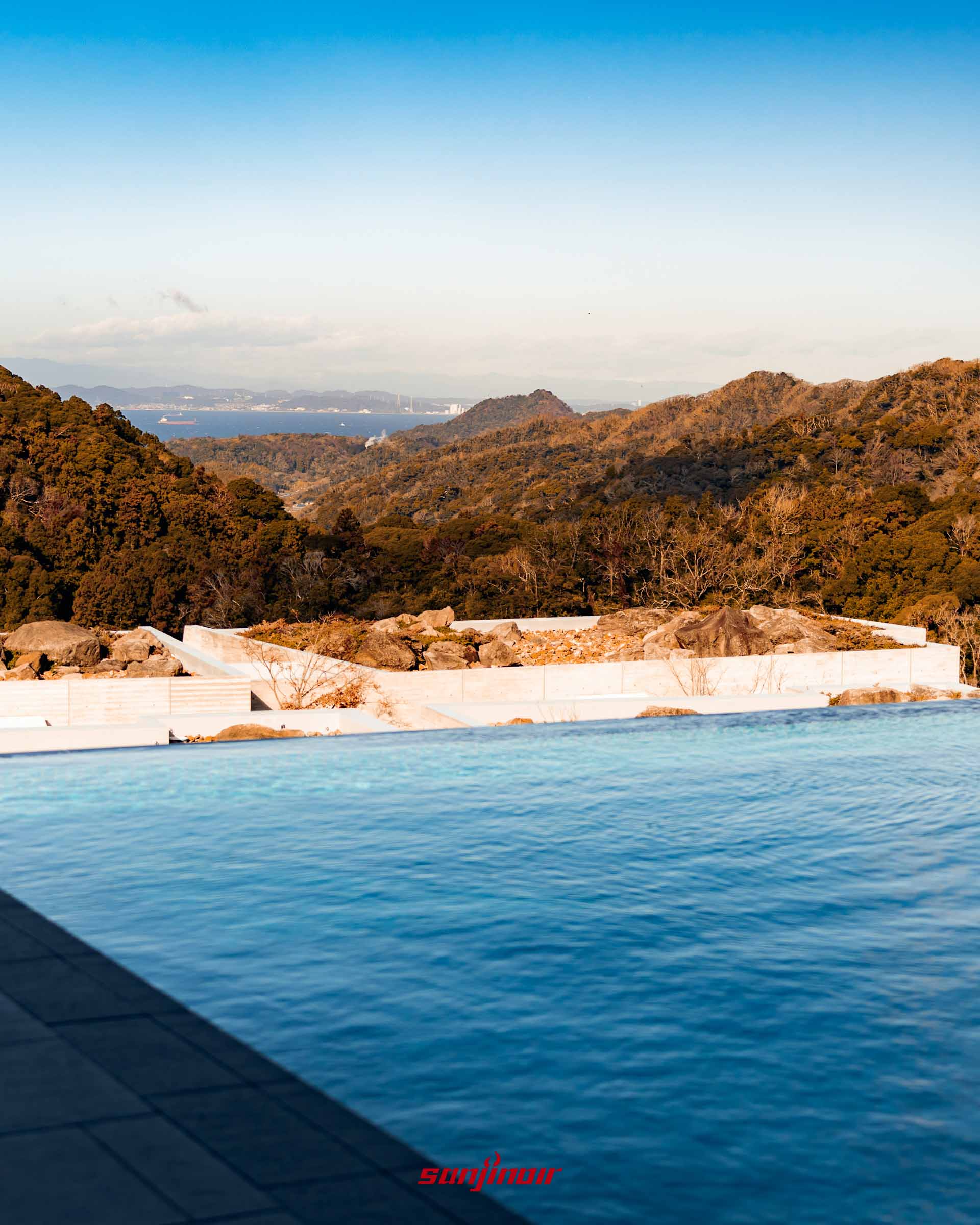 湛藍的無邊際游泳池水面，視覺延伸至遠方的綠色山丘與平靜的東京灣海面，天空呈現晴朗的漸層藍。