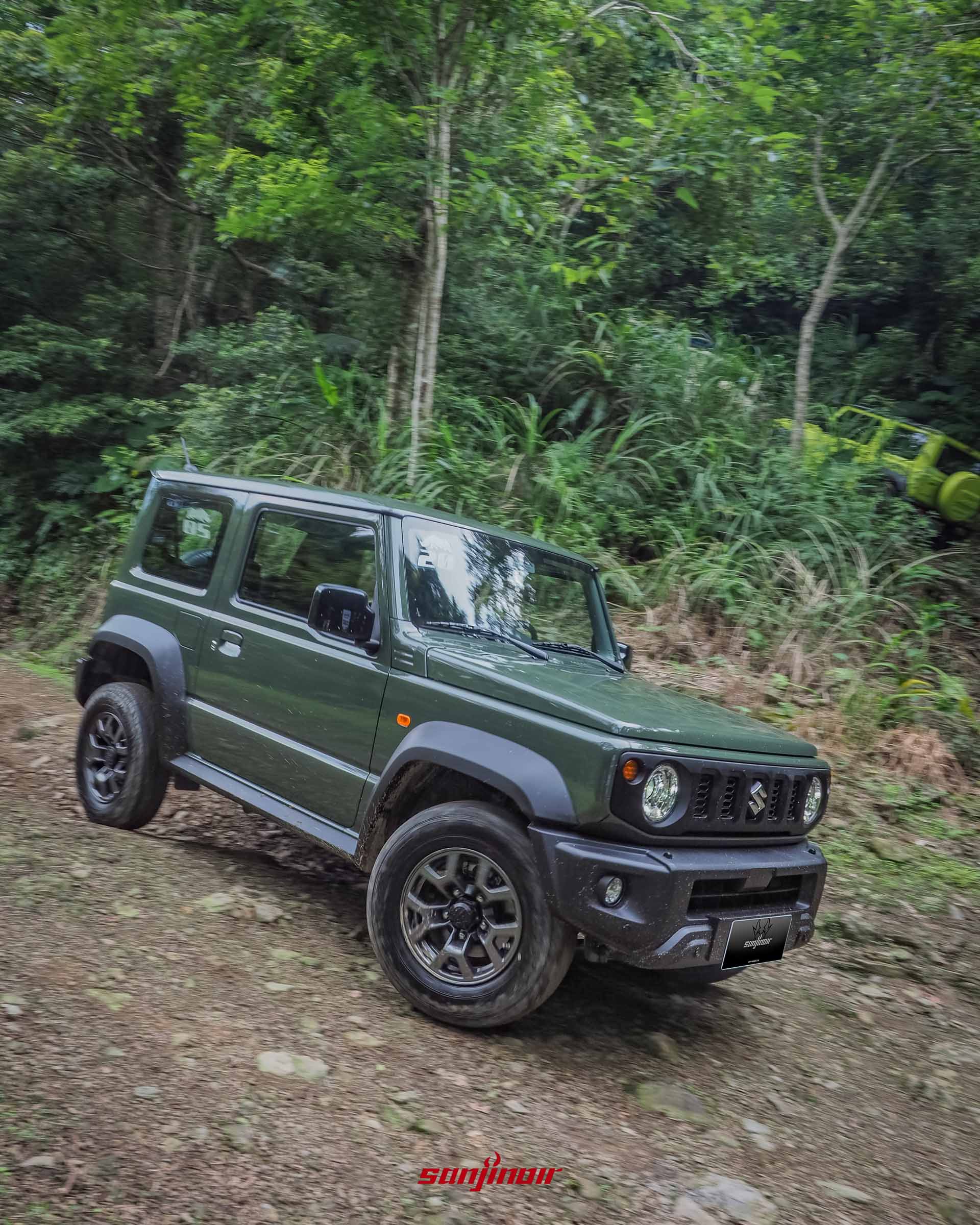 開啟頭燈的軍綠色SUZUKI Jimny正在跨越崎嶇泥濘林道的正面照片。