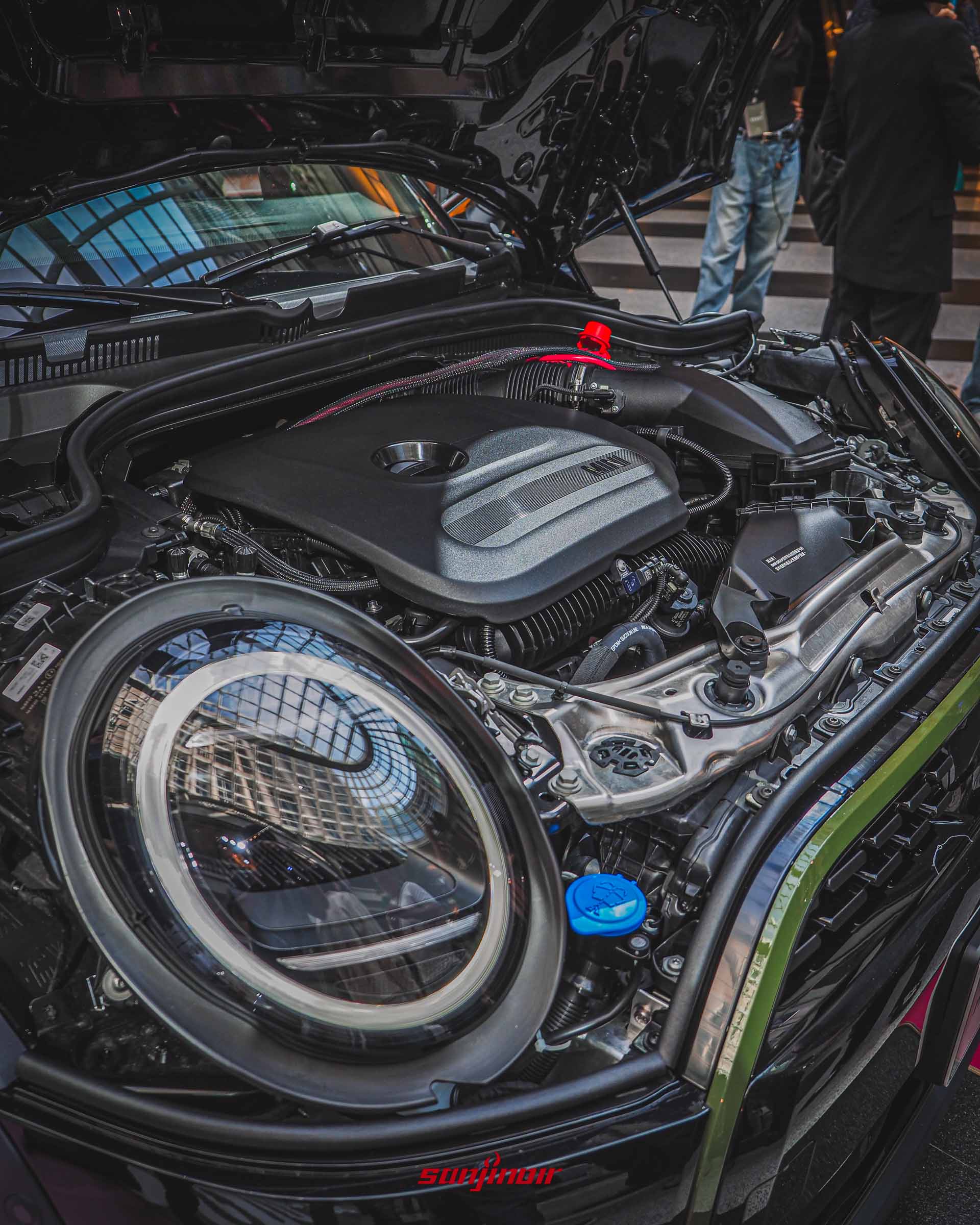 打開引擎蓋的MINI車頭特寫，展示出內部緊湊的引擎室結構與黑色的引擎護蓋，前景是帶有環形日行燈的圓形LED頭燈。