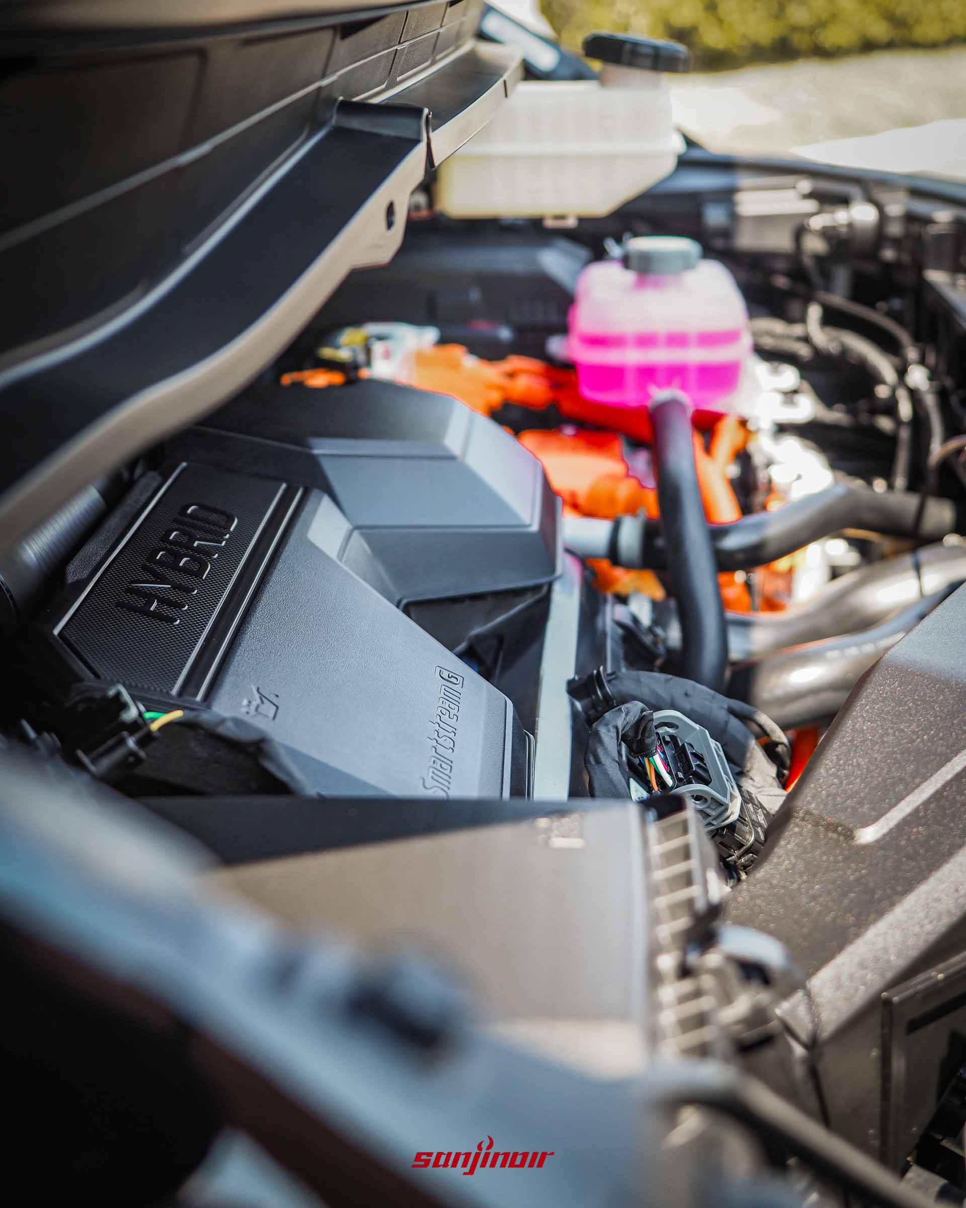 打開引擎蓋特寫 Hyundai Staria 的 1.6 Turbo Hybrid 引擎室,可見帶有 HYBRID 字樣的黑色引擎飾蓋與橘色高壓線束。