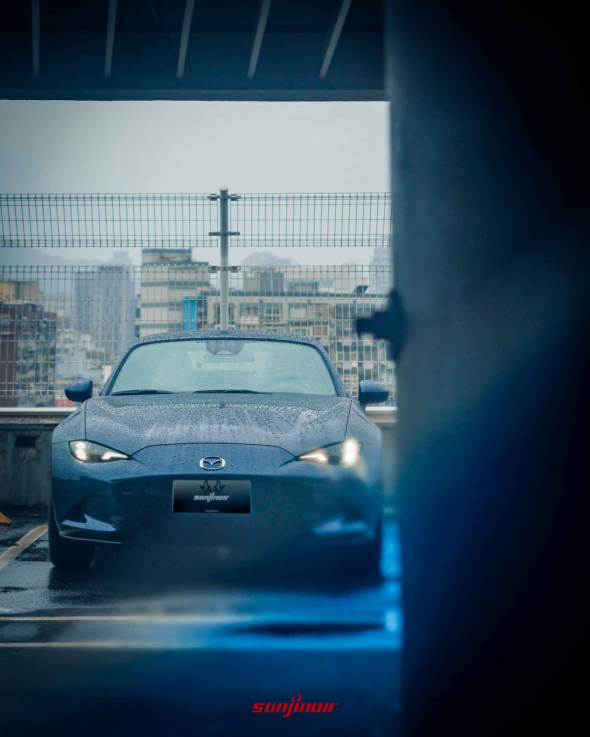 從柱子後方窺視雨中的深藍色 Mazda MX-5 RF 車頭,帶有神祕且靜謐的視覺氛圍。