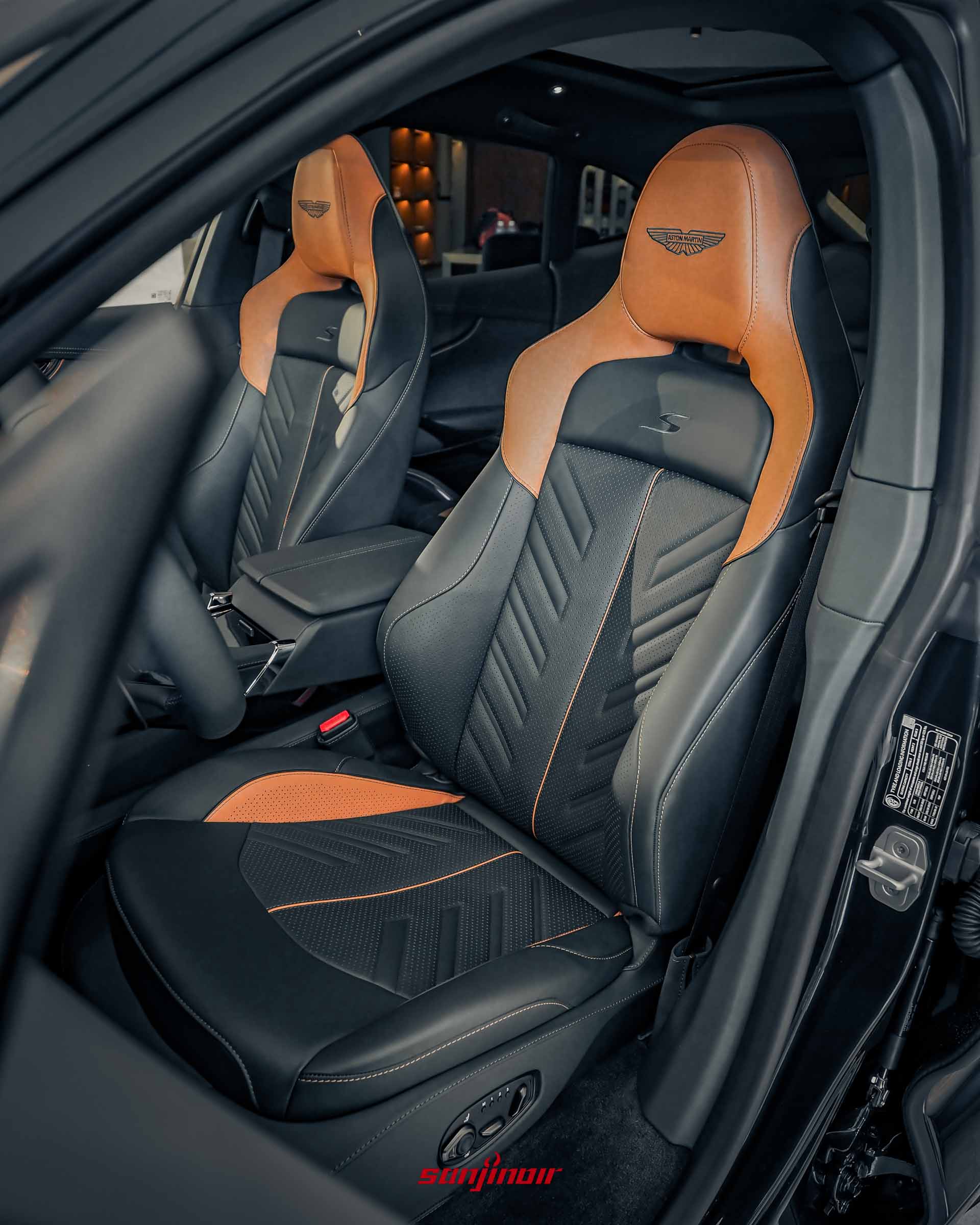 Aston Martin DBX S雙前座跑車座椅特寫，座椅採用深色皮革搭配橘色對比區塊與縫線，椅面具備幾何打孔造型，椅背繡有S字樣，頭枕處則有Aston Martin廠徽浮雕。