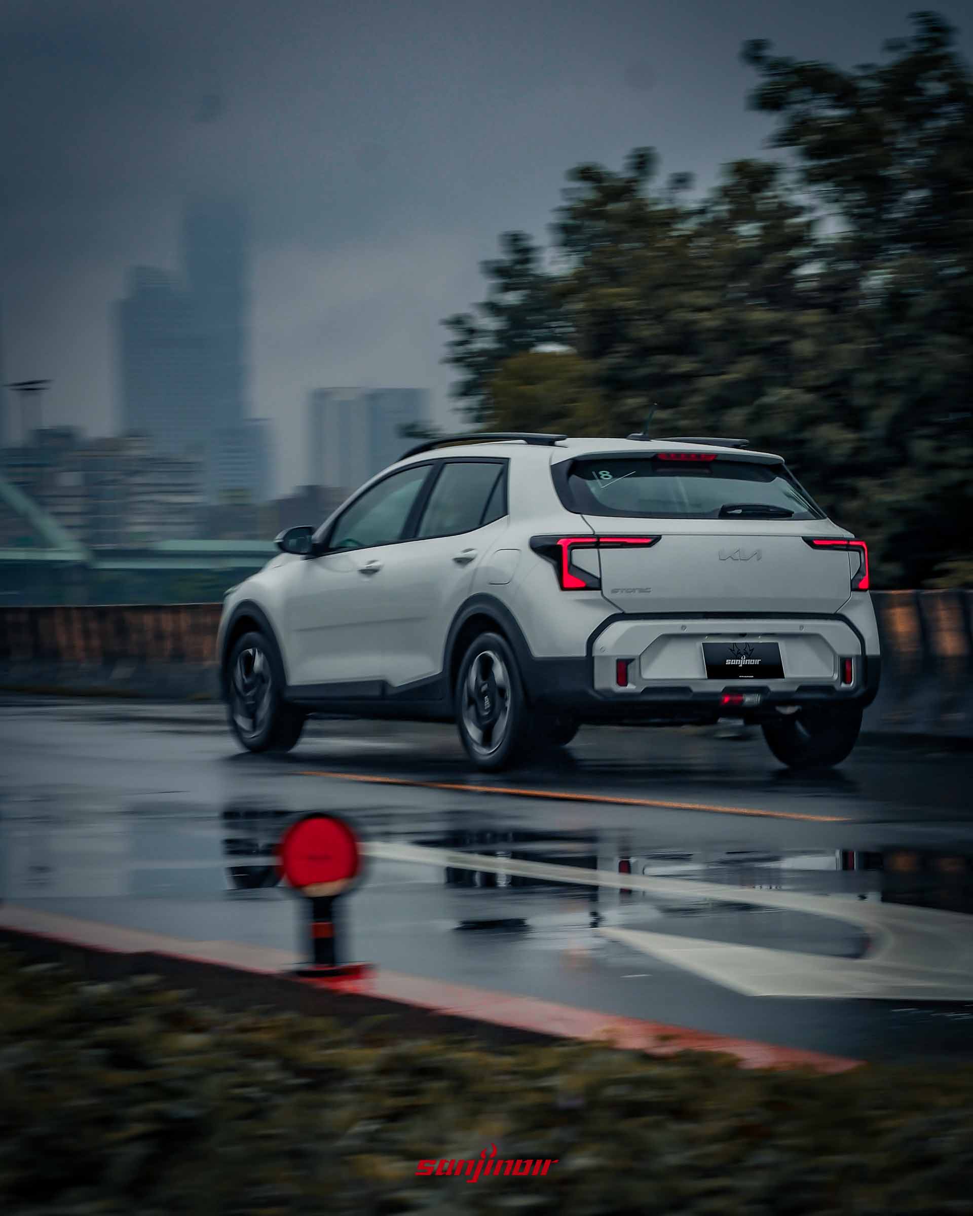 從左後方斜側視角動態捕捉白色 2026 Kia Stonic 行駛於陰雨天濕滑道路,頭尾燈亮起,背景為模糊的城市高樓與樹木。
