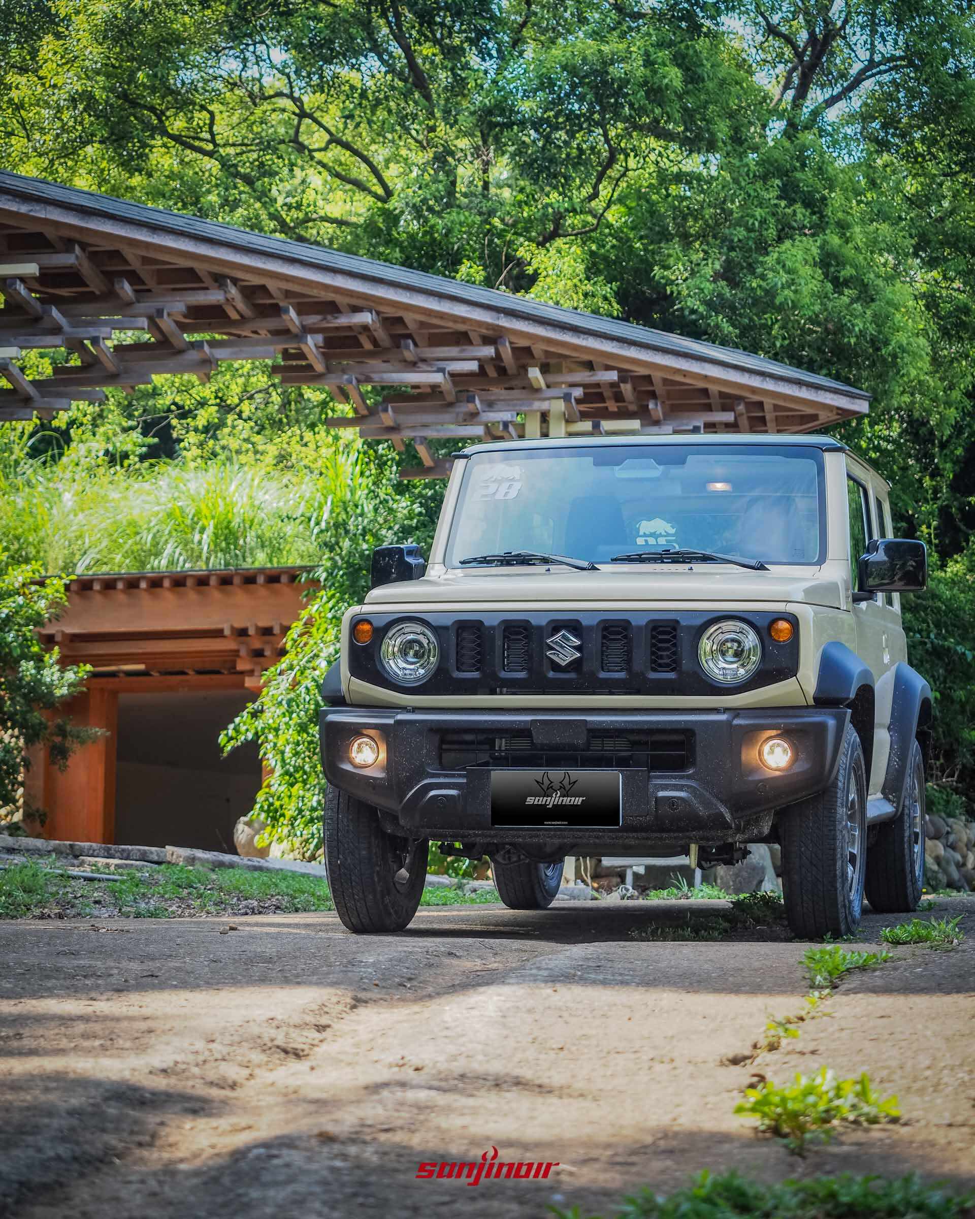 米色SUZUKI Jimny停放於傳統木製建築與綠意盎然背景前的車頭正面照。