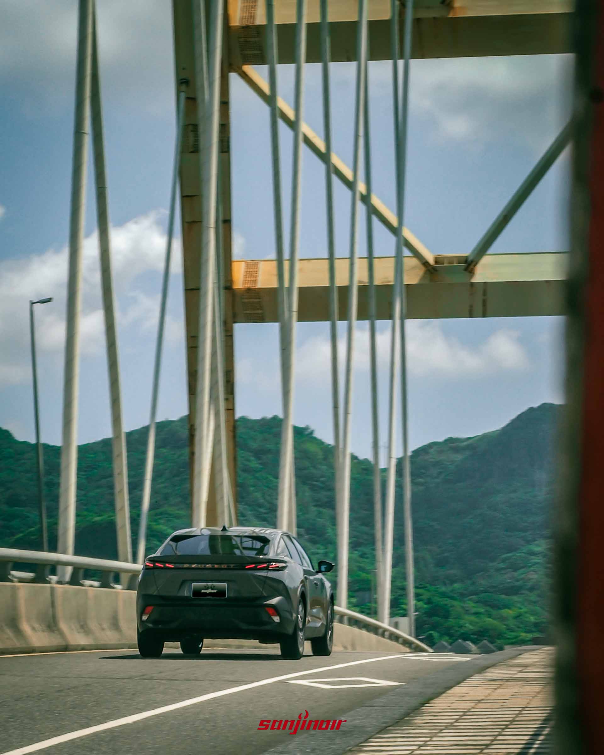 灰色PEUGEOT 408在橋樑上行駛的車尾動態照片,背景為翠綠山丘。