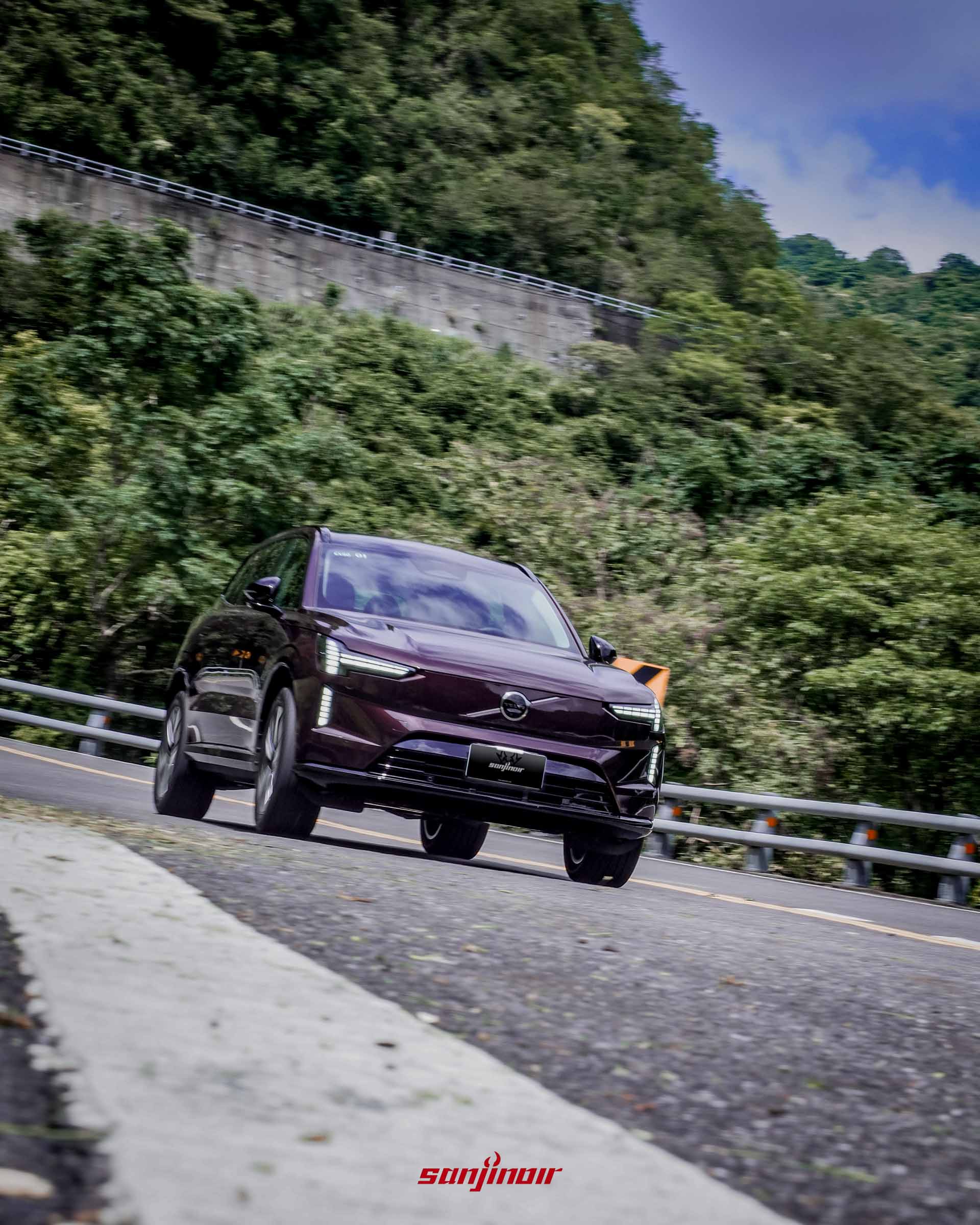 野莓紅Volvo EX90在山區道路過彎時的動態追焦照,車頭朝向畫面左側,背景為綠色植被。