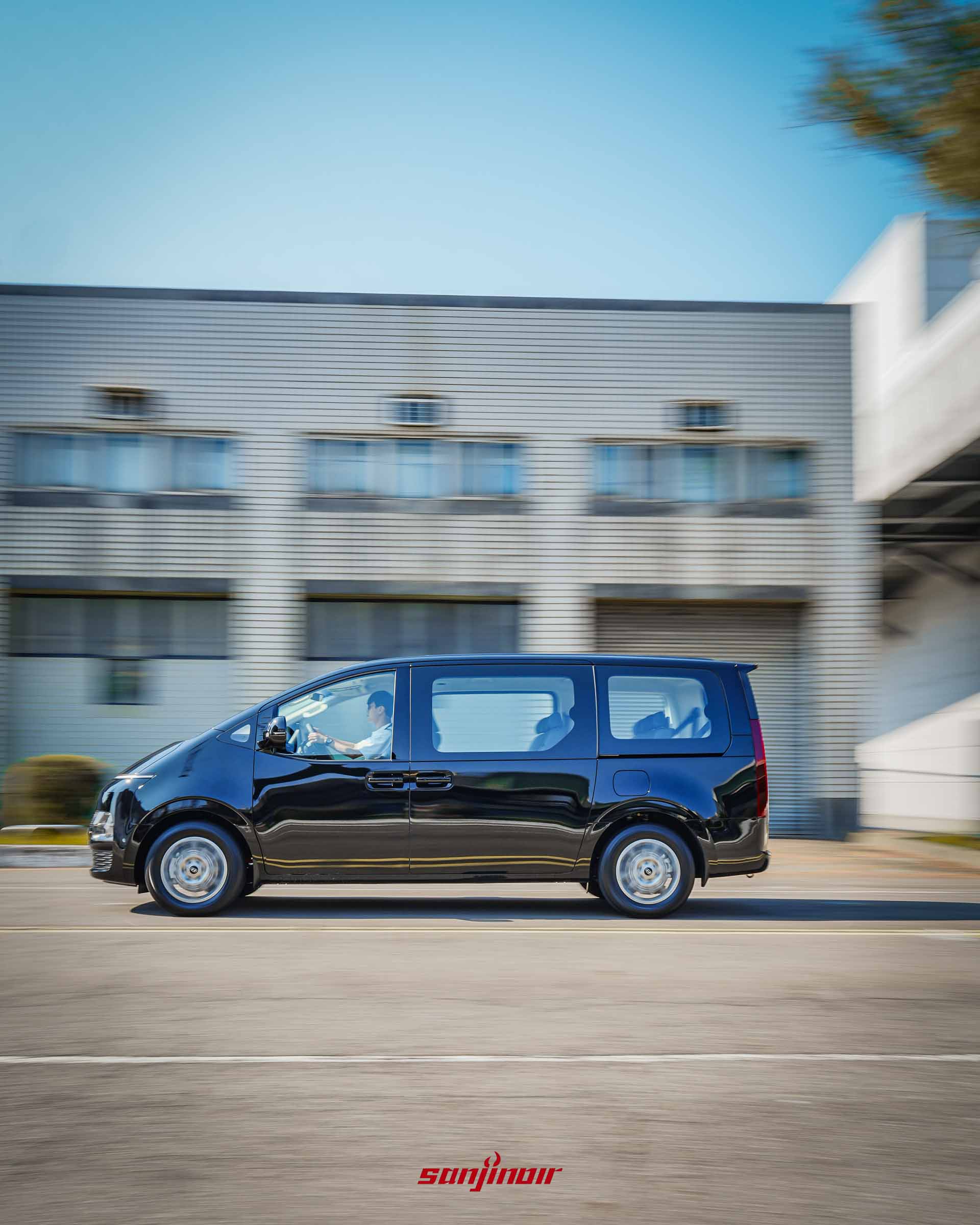 黑色 Hyundai Staria 在道路上行駛的正側面動態追焦攝影,清晰展示其修長的車身比例與大面積平整化車側玻璃。